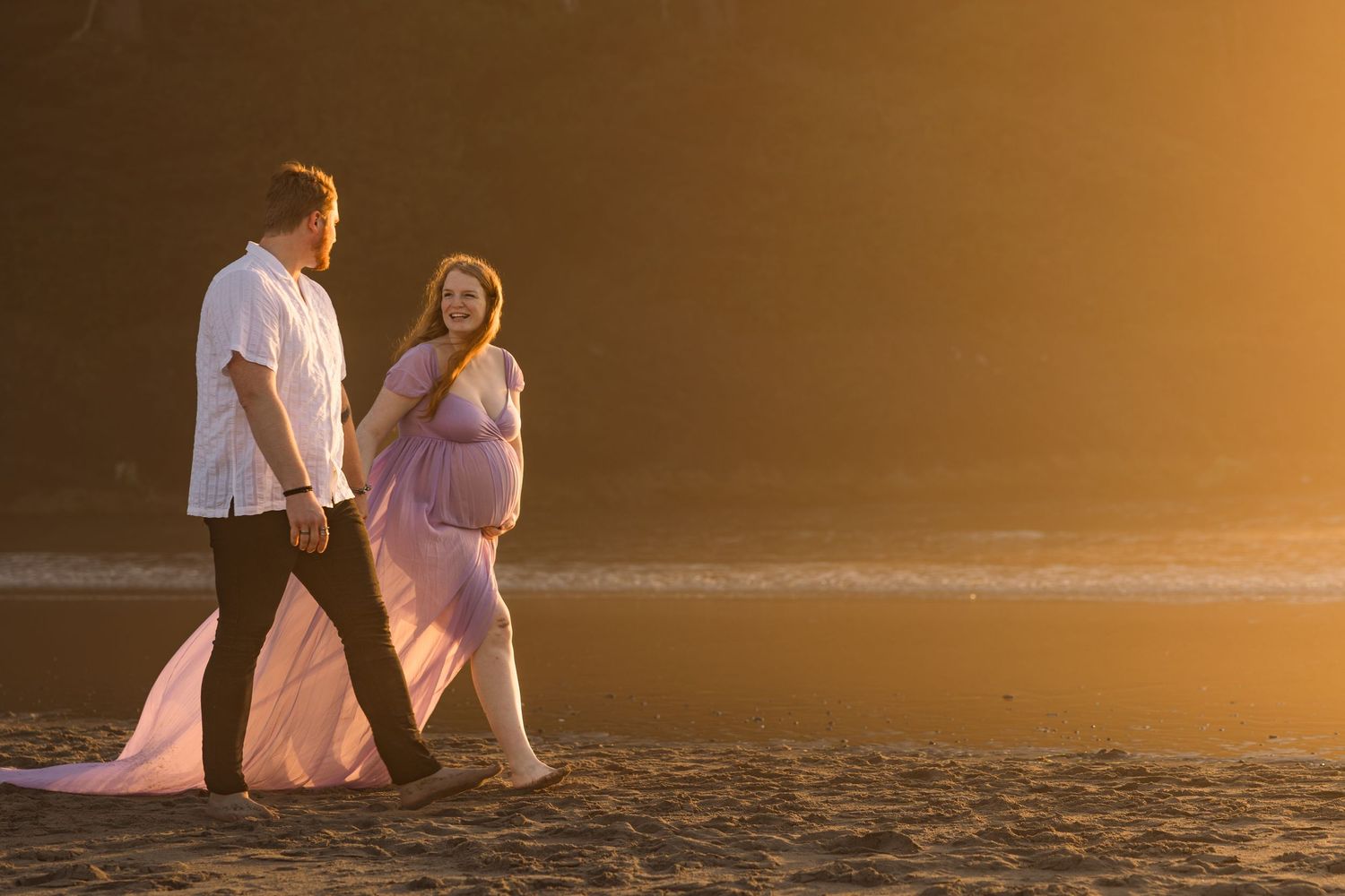 A couple walking along the beach at sunset while silhouetted against a golden sky.