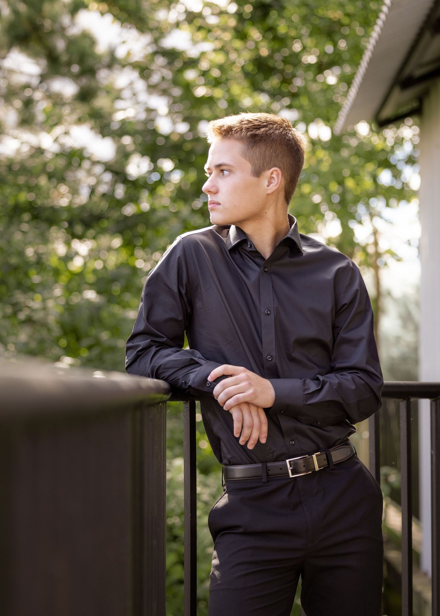 Senior in formal black attire leaning on railing with trees in background during a photo session taken by Salem, Oregon senior photographer.