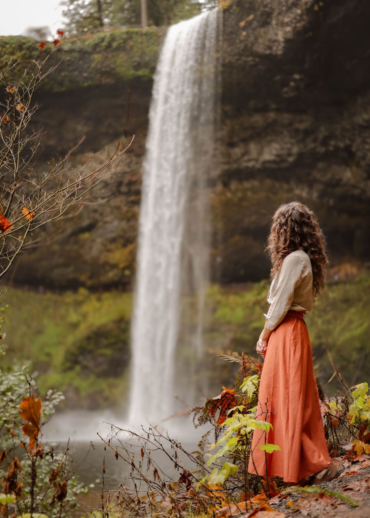 A person in an orange skirt stands near a tall cascading waterfall surrounded by autumn foliage and rocky cliffs.