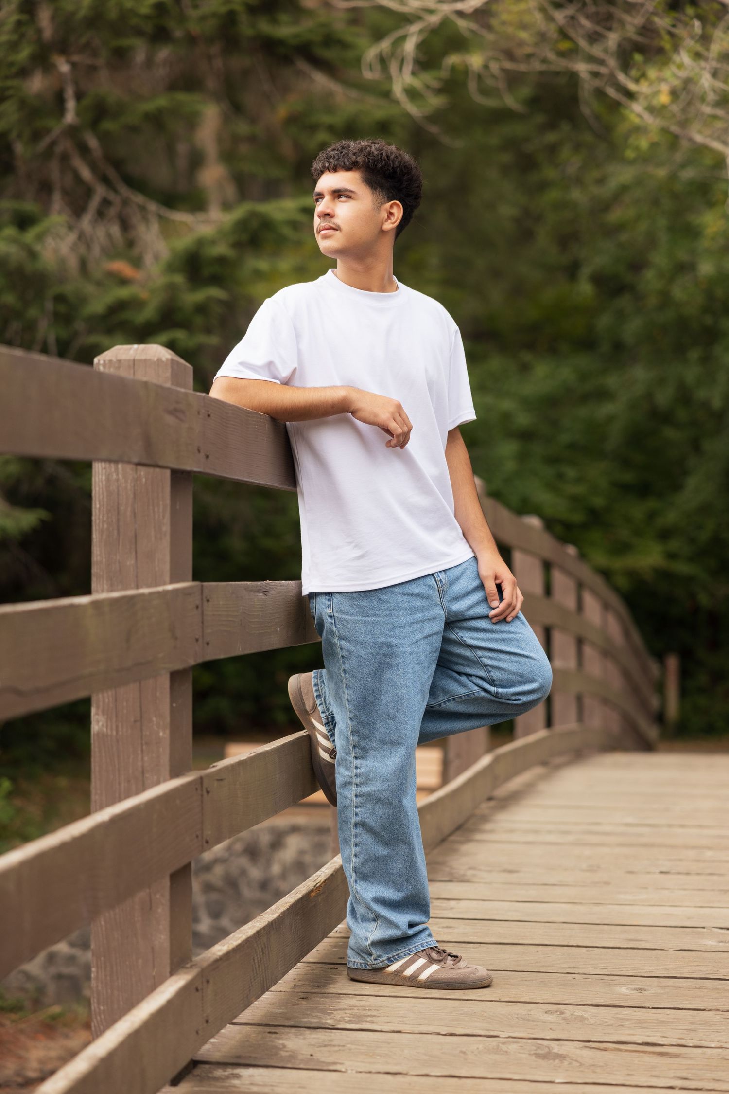 Young man in white t-shirt and jeans leaning on wooden bridge railing surrounded by lush green trees.