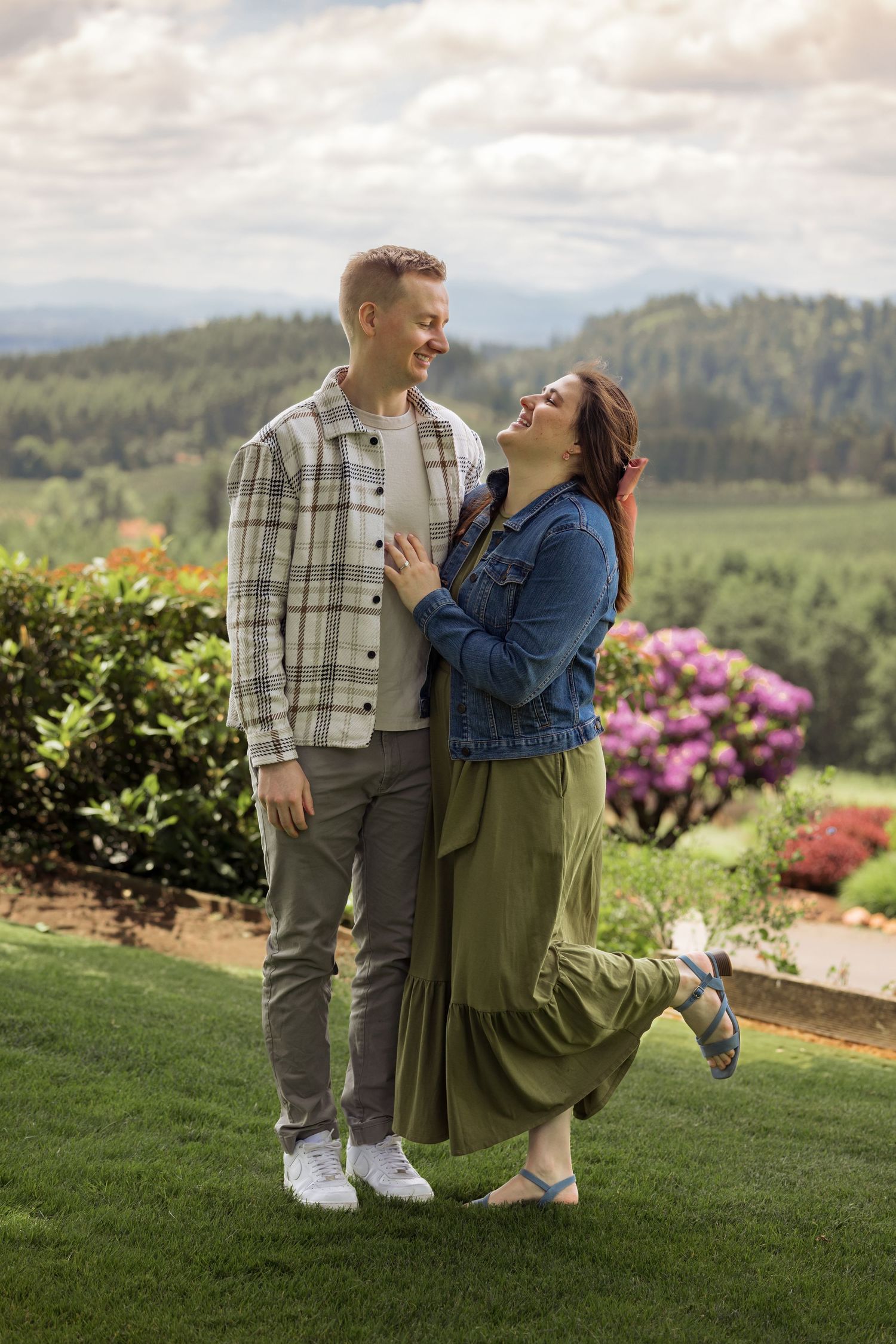A couple laughs together while posing on a scenic overlook with mountains and colorful gardens behind them.