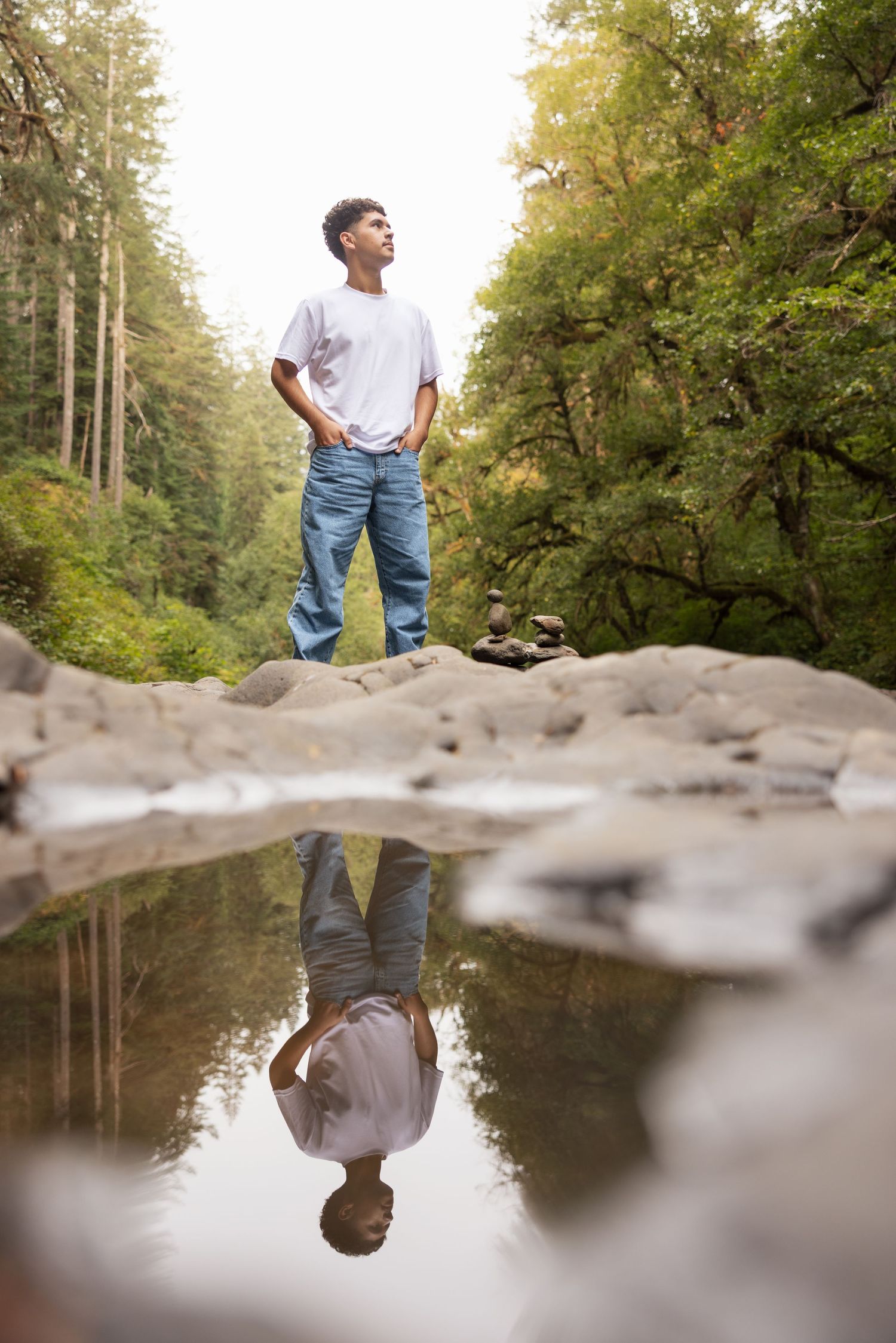 Young man standing on rocks by a reflective forest pool, hands in pockets, gazing upward surrounded by lush green trees.