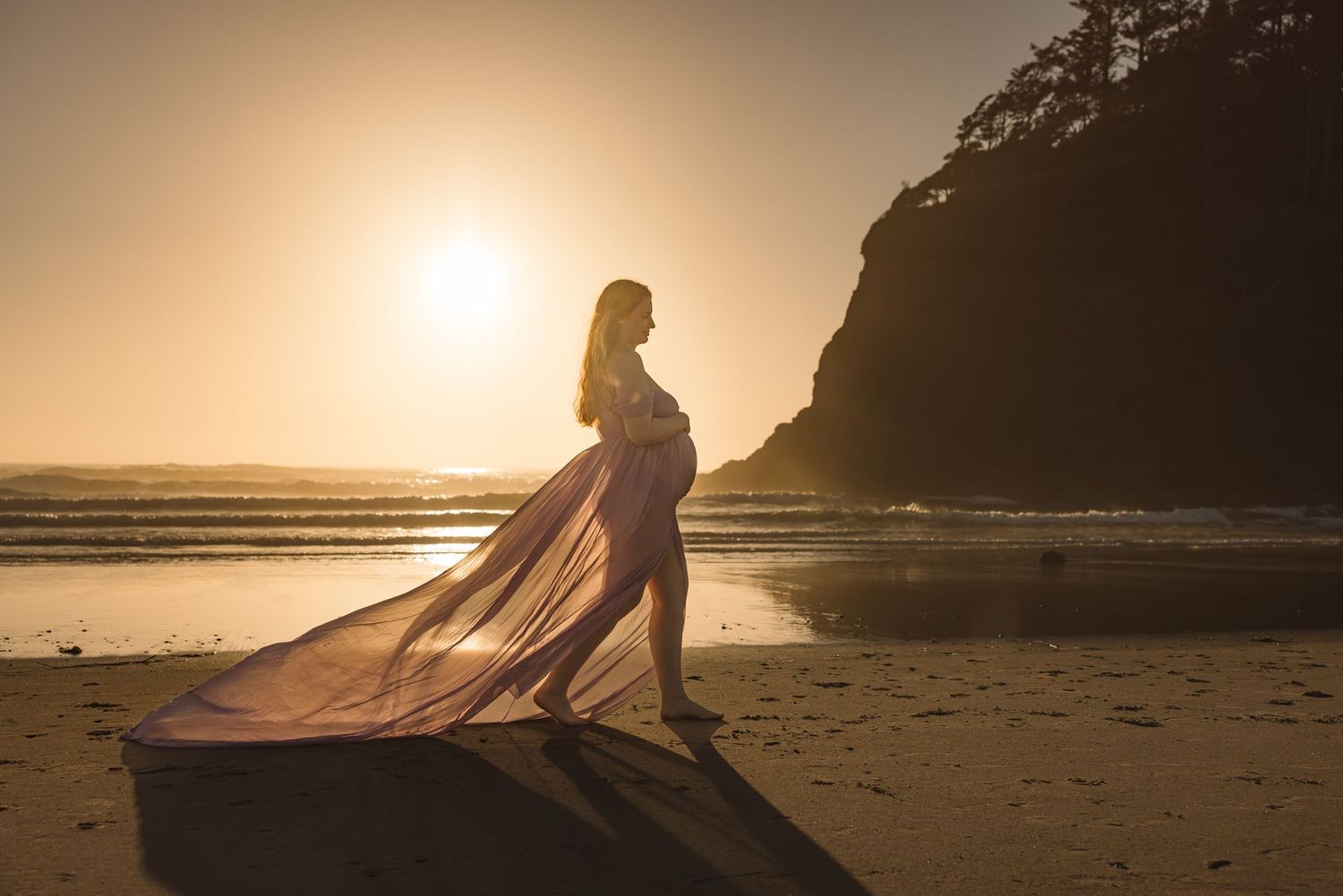Silhouette stands on beach at sunset with flowing dress fabric catching the golden light near coastal cliffs.