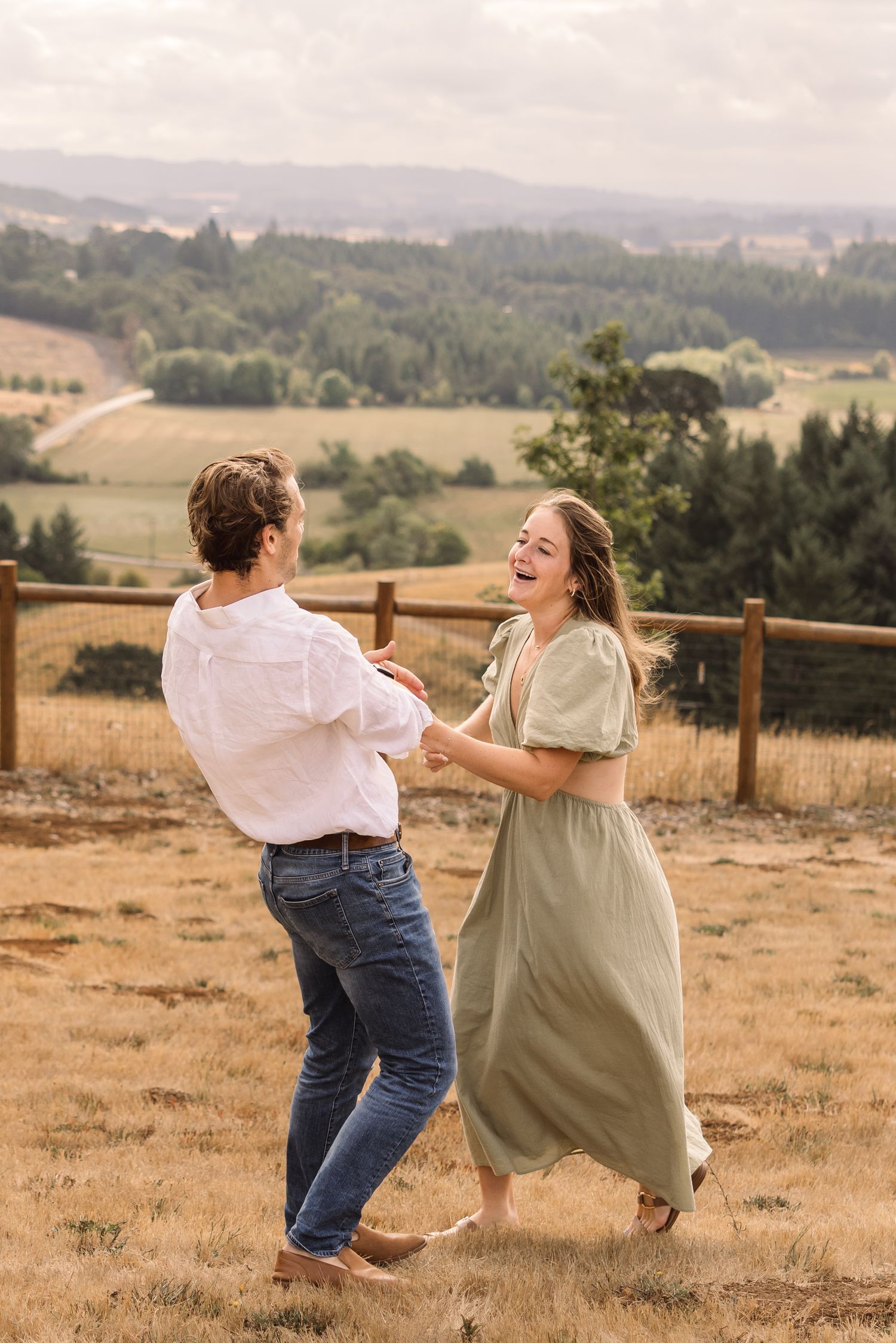 A couple dances and laughs together on a hilltop overlooking rolling countryside hills during golden hour.
