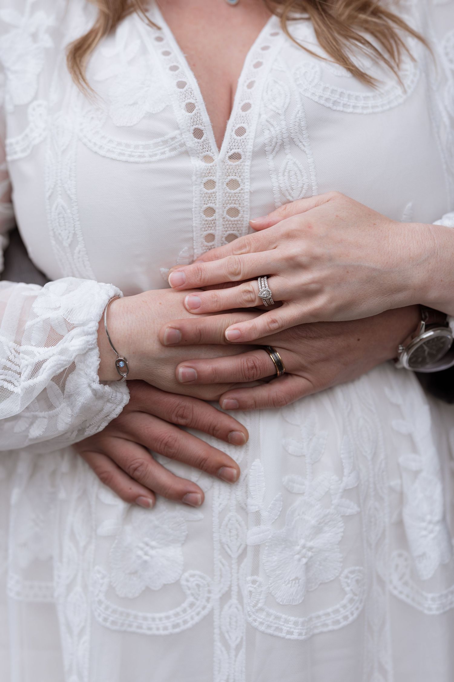 Couple holding hands showing wedding rings, woman in white lace dress with man's arm wrapped around her waist.