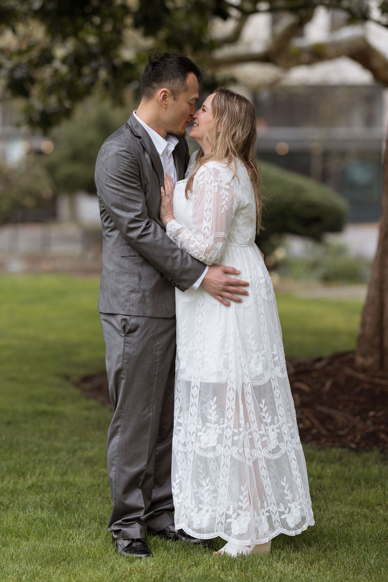 Couple sharing a romantic kiss outdoors, bride in white lace gown and groom in gray suit on green lawn.