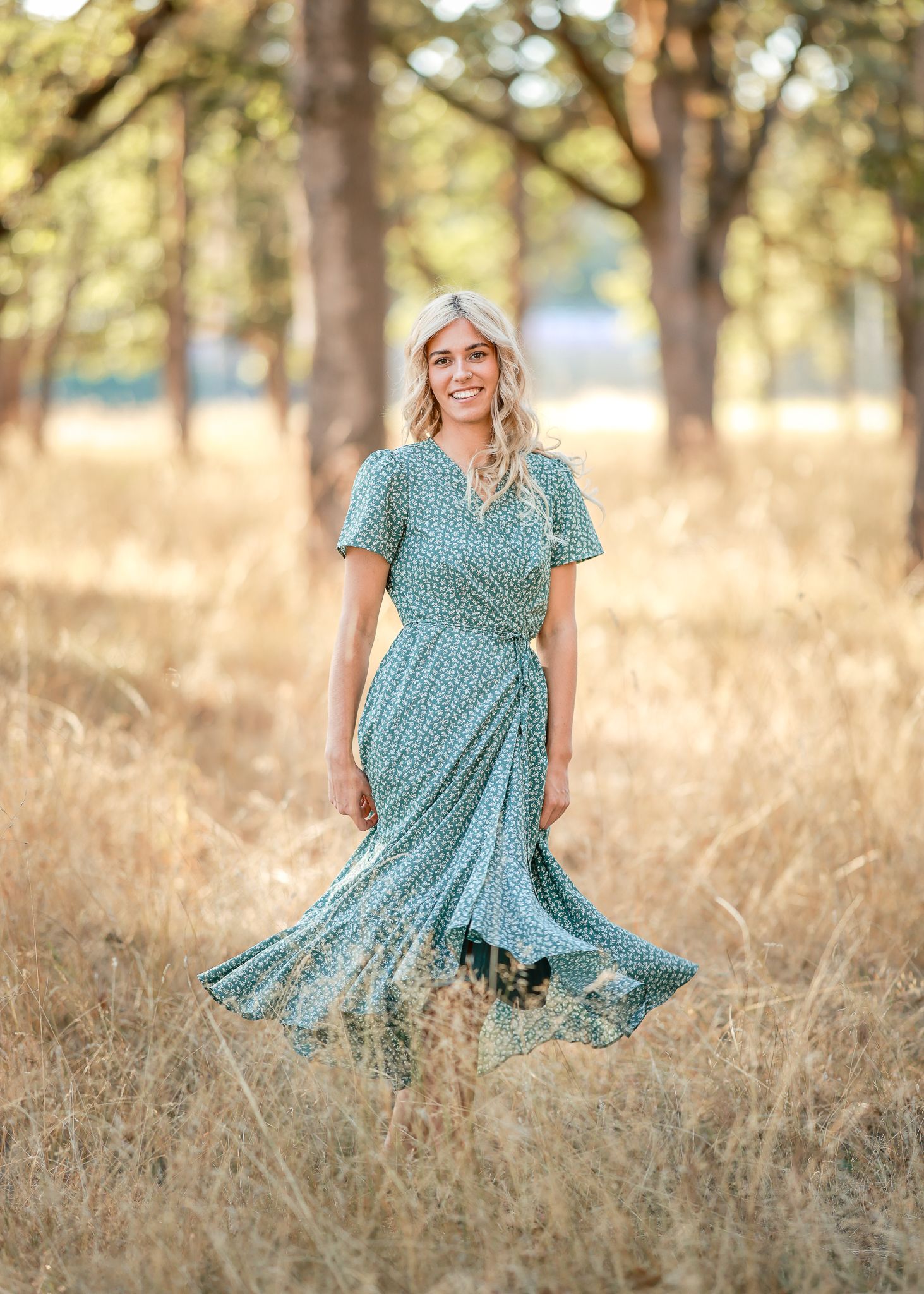 Woman in teal dress twirls through golden field at sunset.