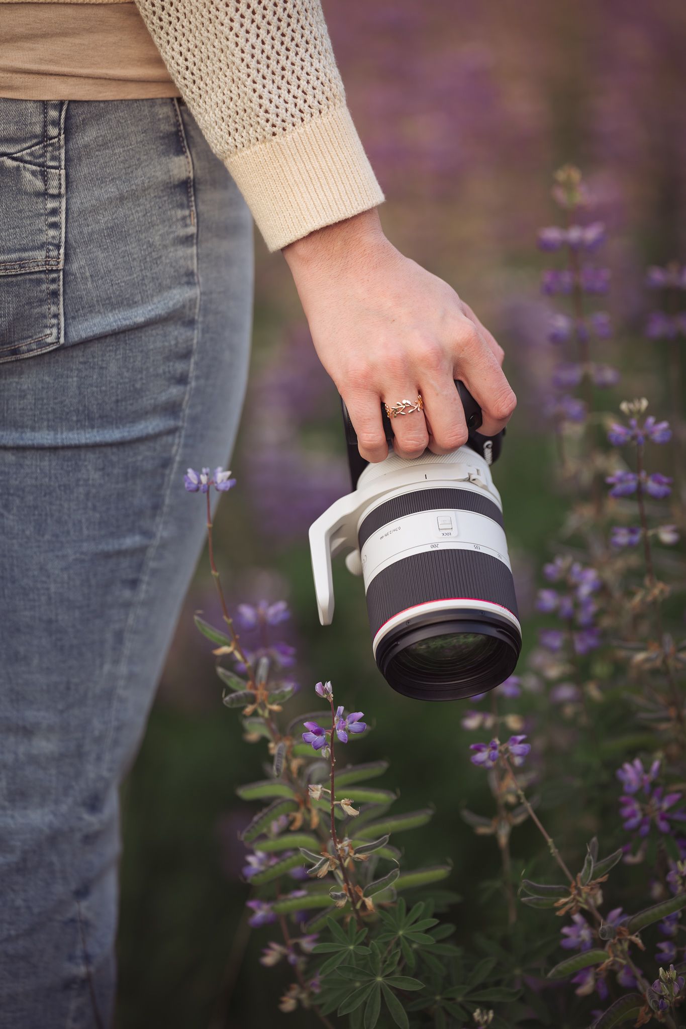 Hand wearing engagement ring holds camera lens among purple wildflowers.