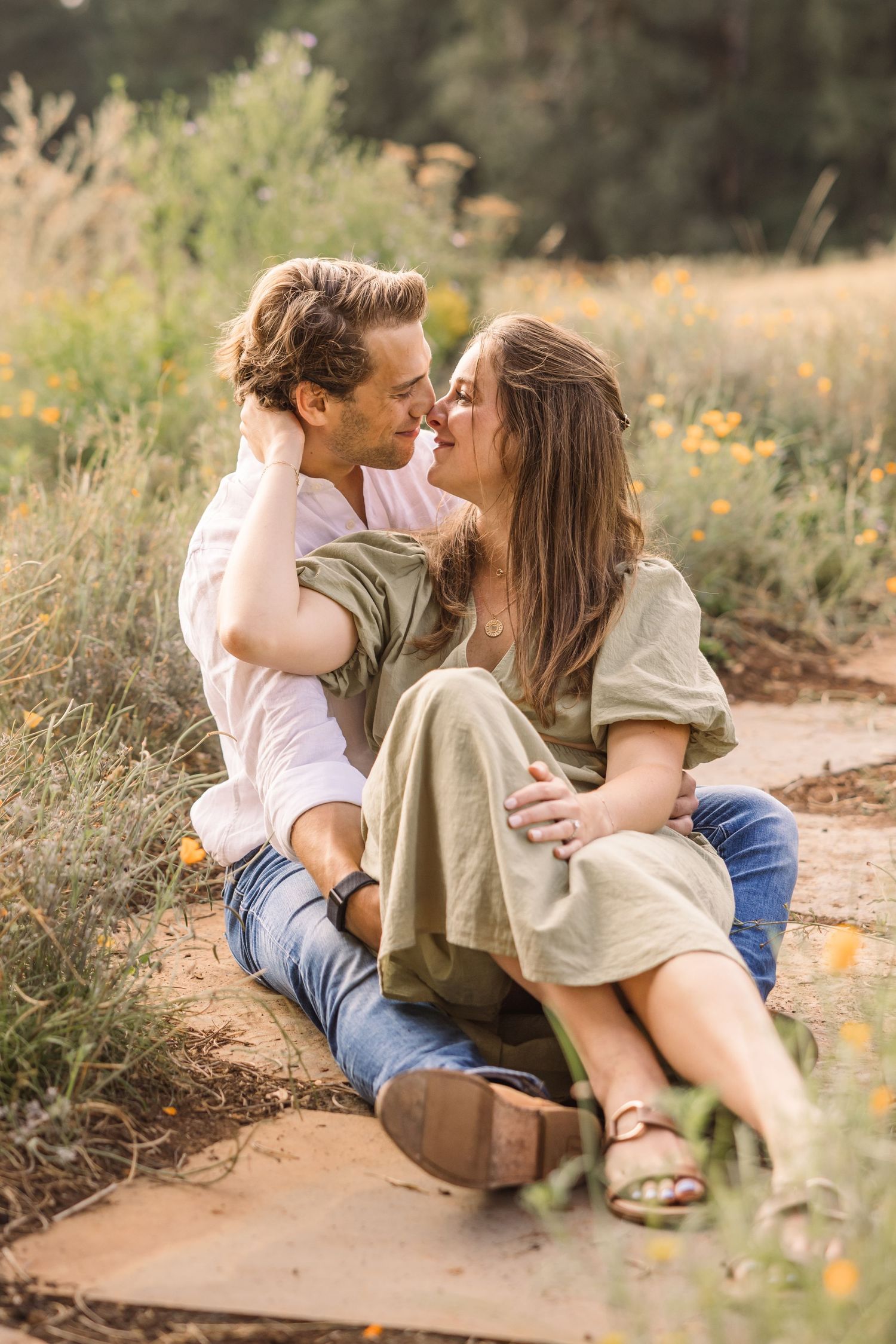 A romantic couple shares intimate moments while sitting on a dirt path surrounded by wildflowers and natural scenery.