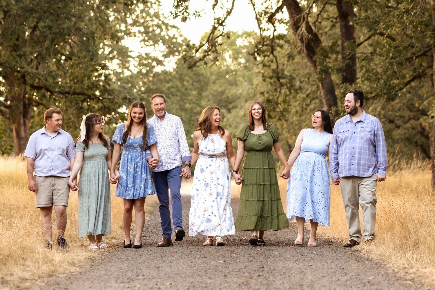 Eight family members walk hand-in-hand down a tree-lined dirt path, dressed in coordinated pastel and earth-tone outfits.