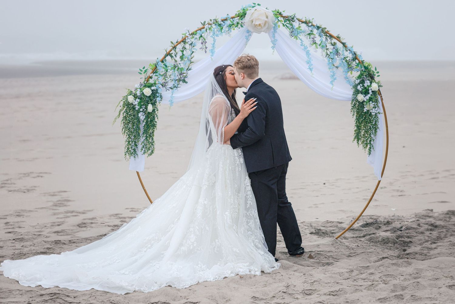 Bride and groom kissing under a floral hoop arch on a sandy beach during their romantic outdoor wedding ceremony.