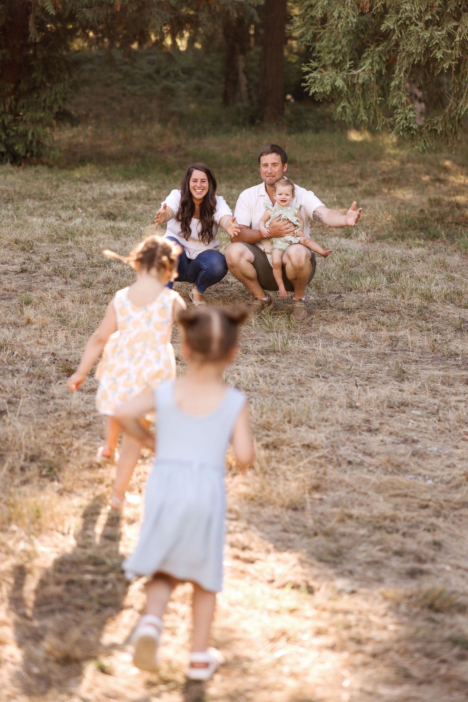 Happy family with two young girls running toward parents crouching in a sunny outdoor park setting.