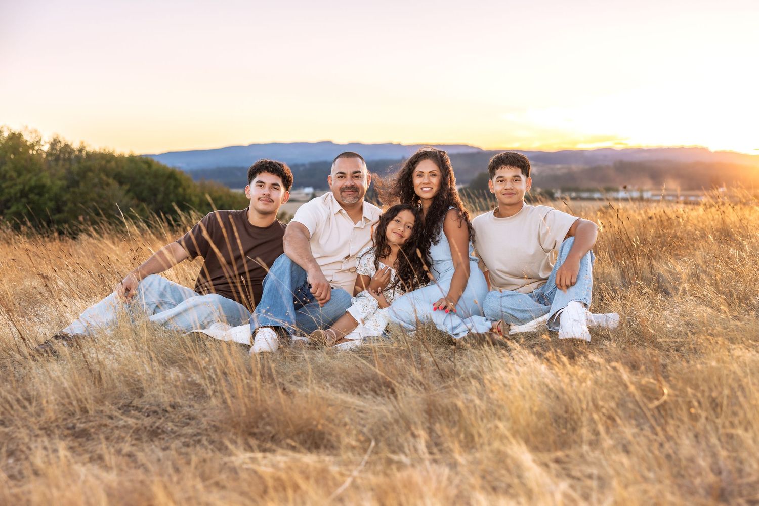 Family of five sitting in golden field at sunset with mountain views in warm natural light.