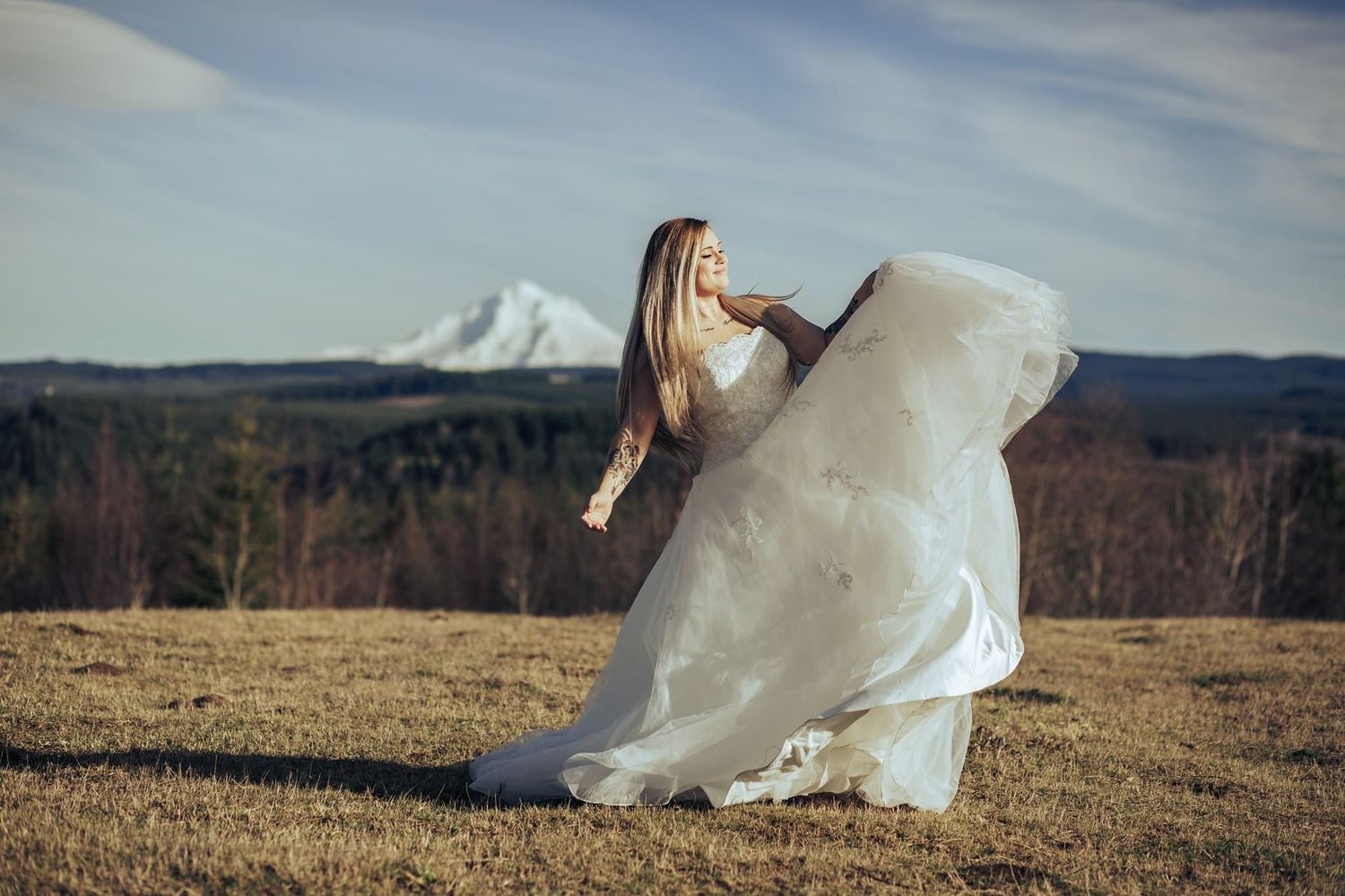 Bride in flowing white wedding dress twirling in an open field with mountain backdrop and blue sky.