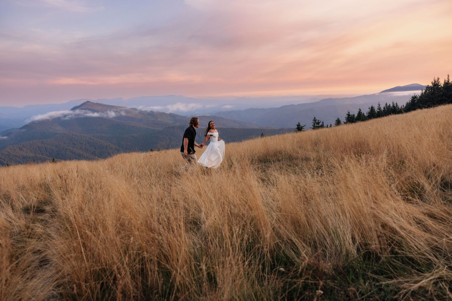 Couple in wedding attire walking through golden meadow grass at sunset with misty mountain views.