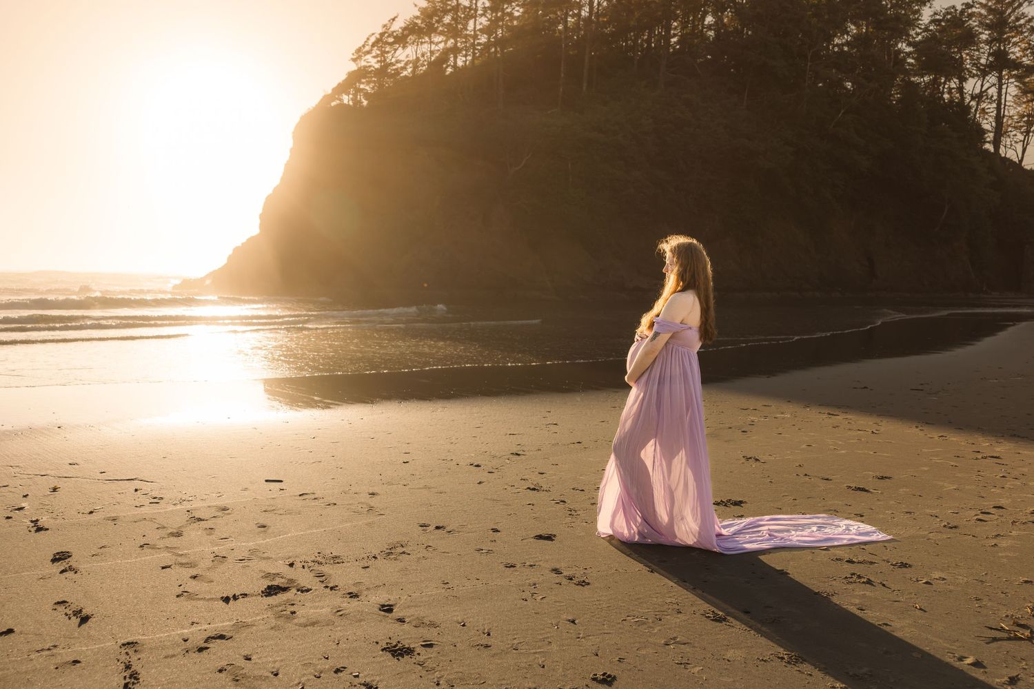 A person in a flowing pink dress stands on a beach at sunset with cliffs and ocean waves in the background.