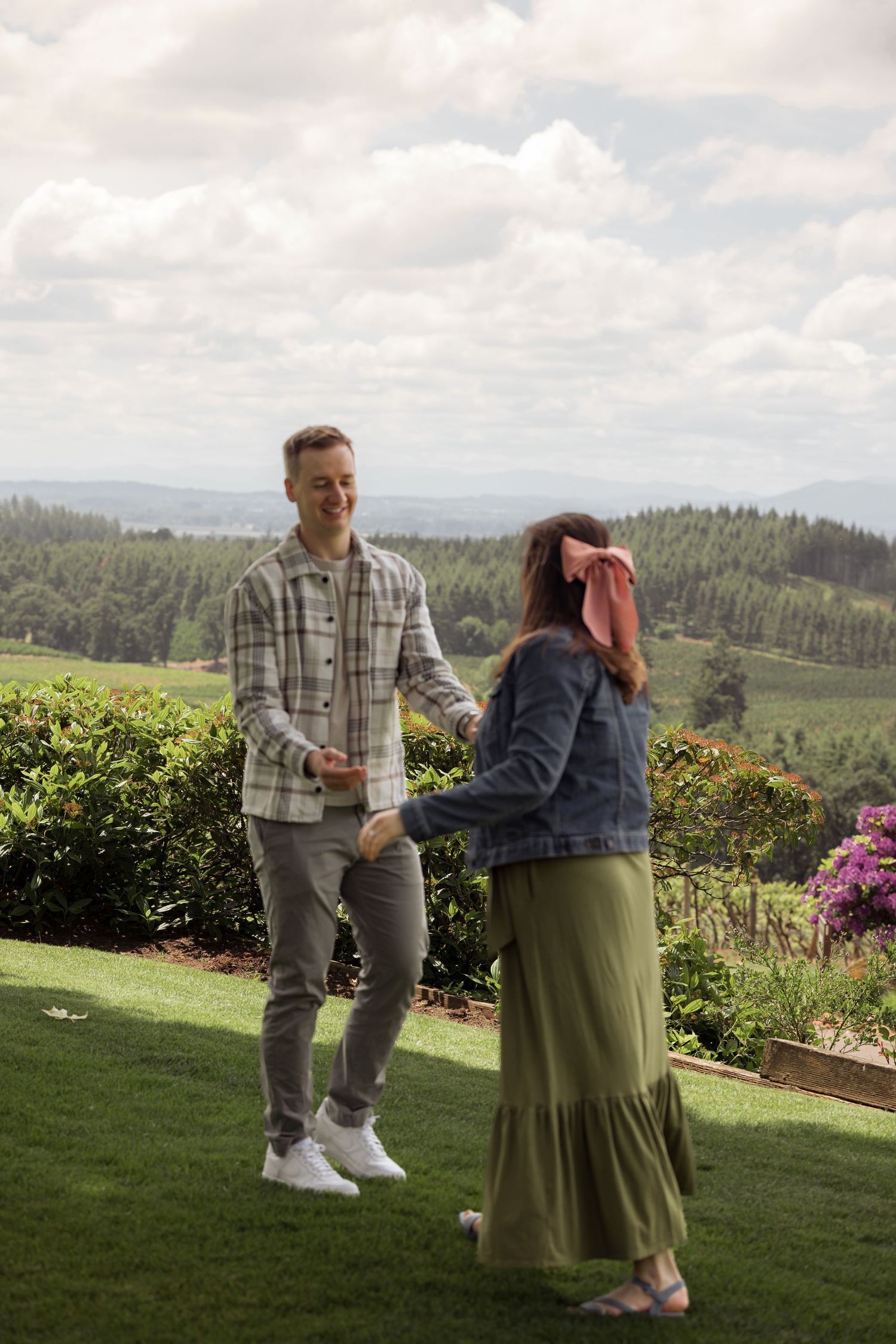 Two people laughing and conversing while standing on a grassy hilltop garden overlooking mountains and forest scenery.