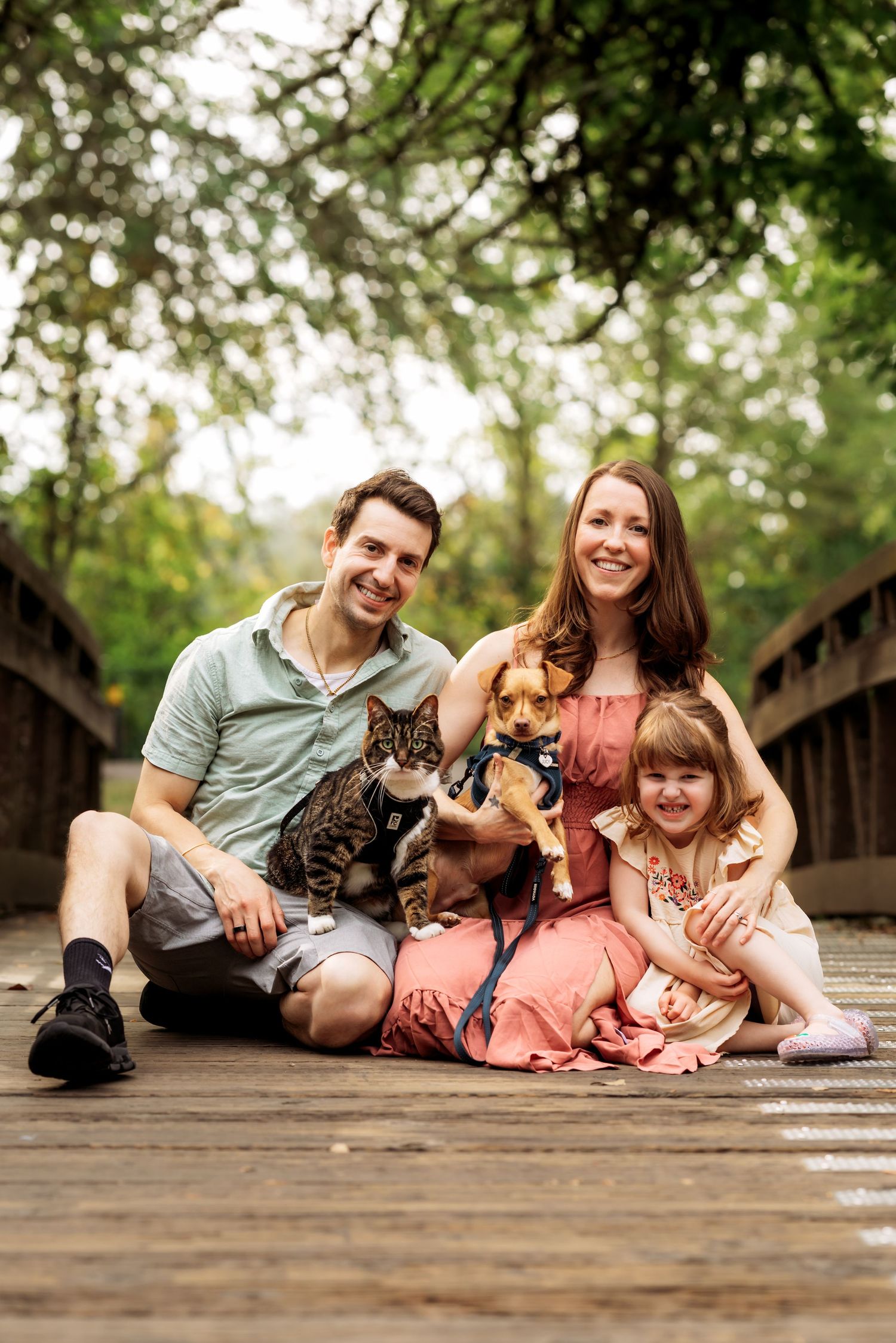 Family of three with a cat and dog posing on a wooden bridge surrounded by lush green trees.