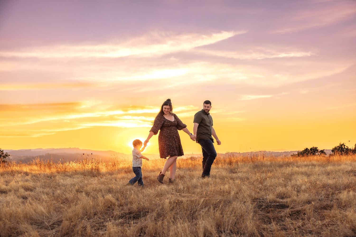 Family of three holding hands walking through golden field at sunset with vibrant pink and purple sky.