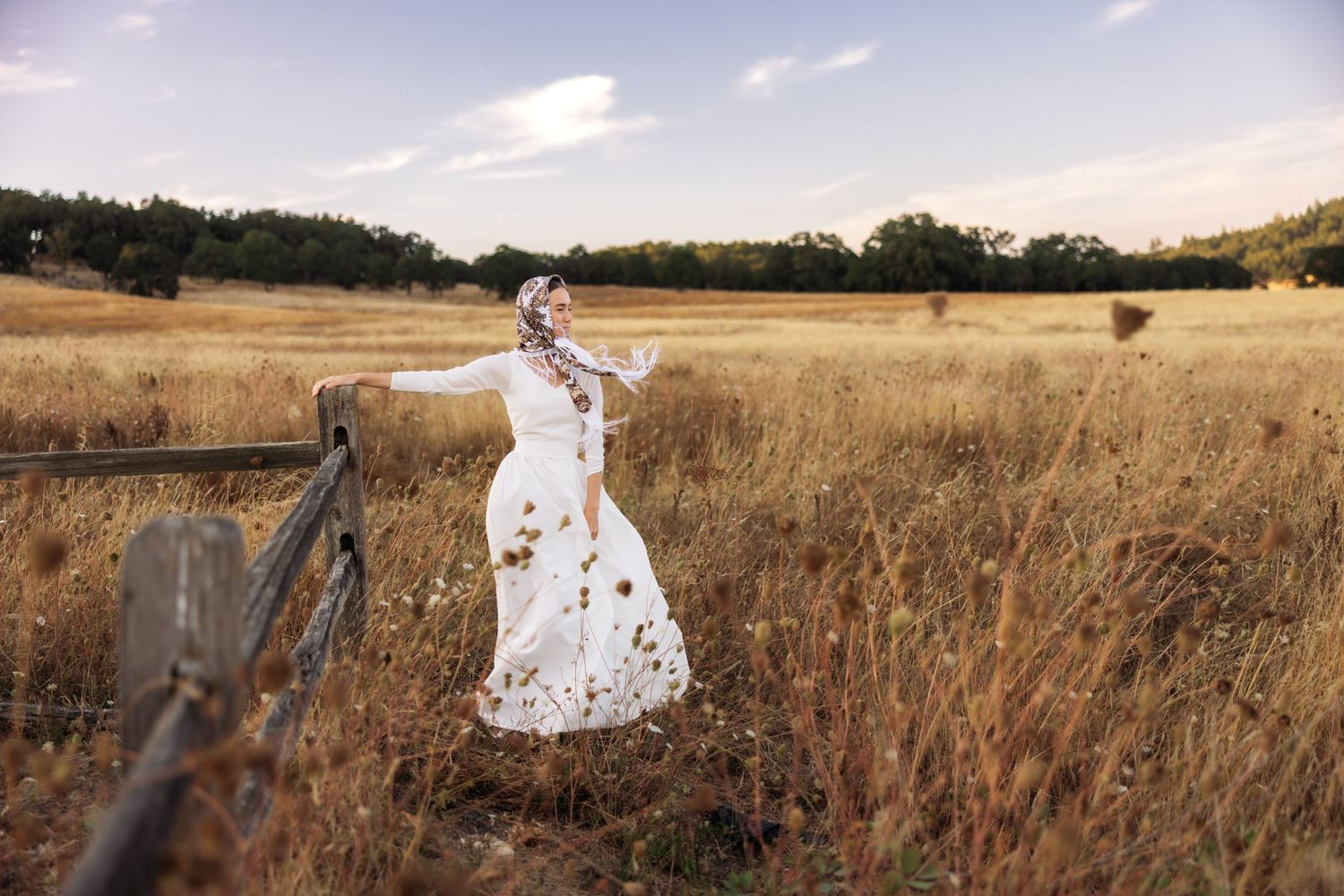 A woman in a white lace dress and floral headscarf stands in a golden field near a wooden fence.