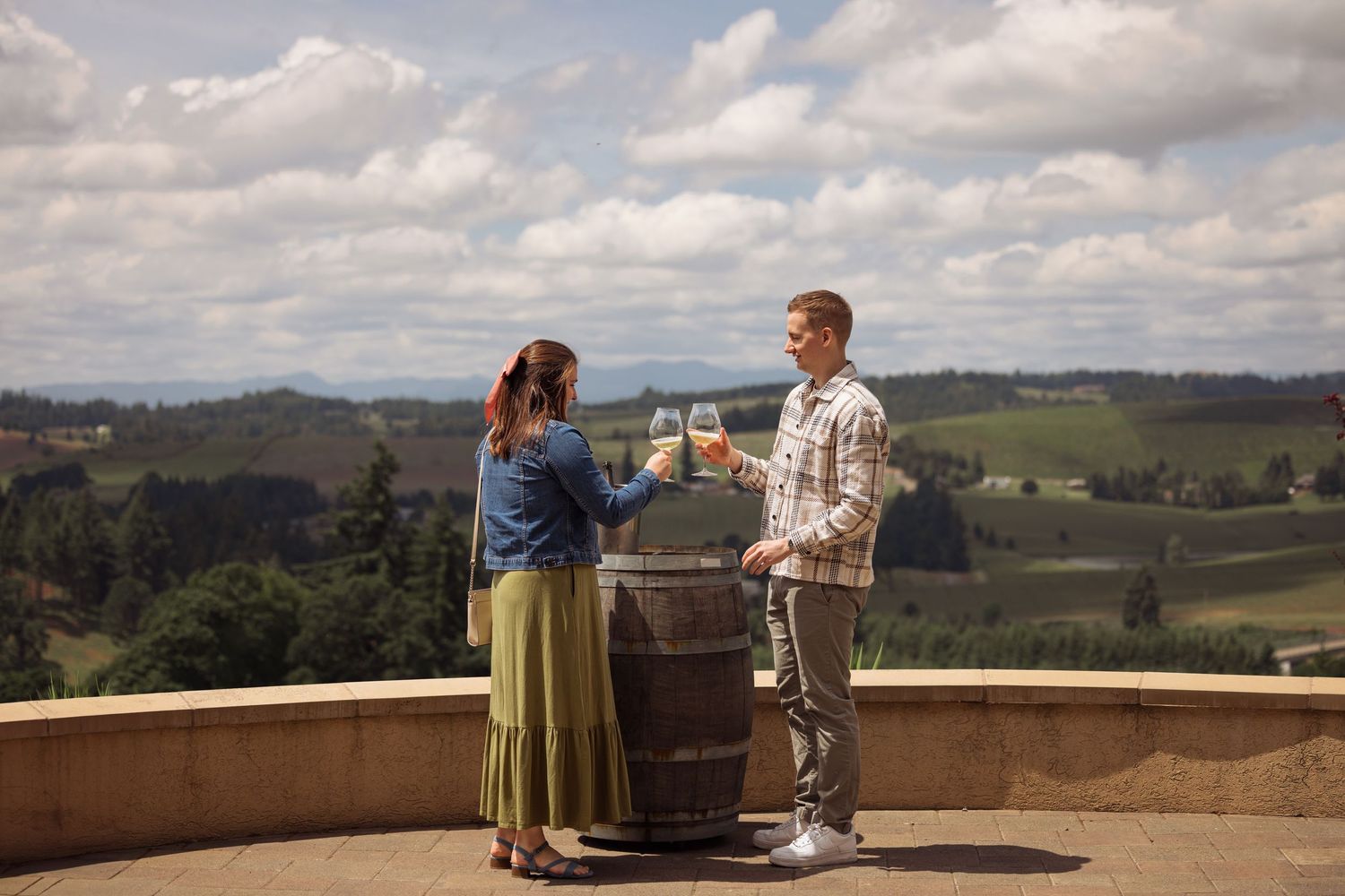 Two people wine tasting at an outdoor terrace overlooking rolling hills and vineyards on a cloudy day.