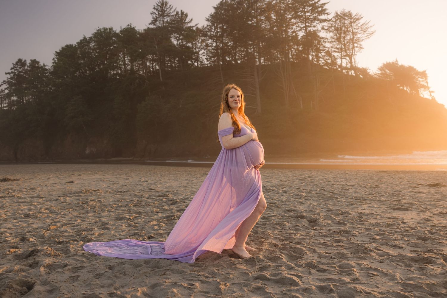 Maternity photo session at sandy beach with flowing violet dress against forested backdrop at golden hour.