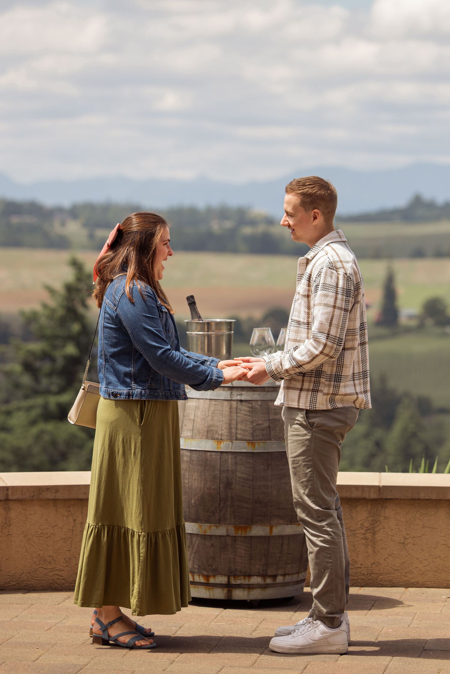 Two people stand next to a wine barrel on a scenic overlook with rolling hills in the background.