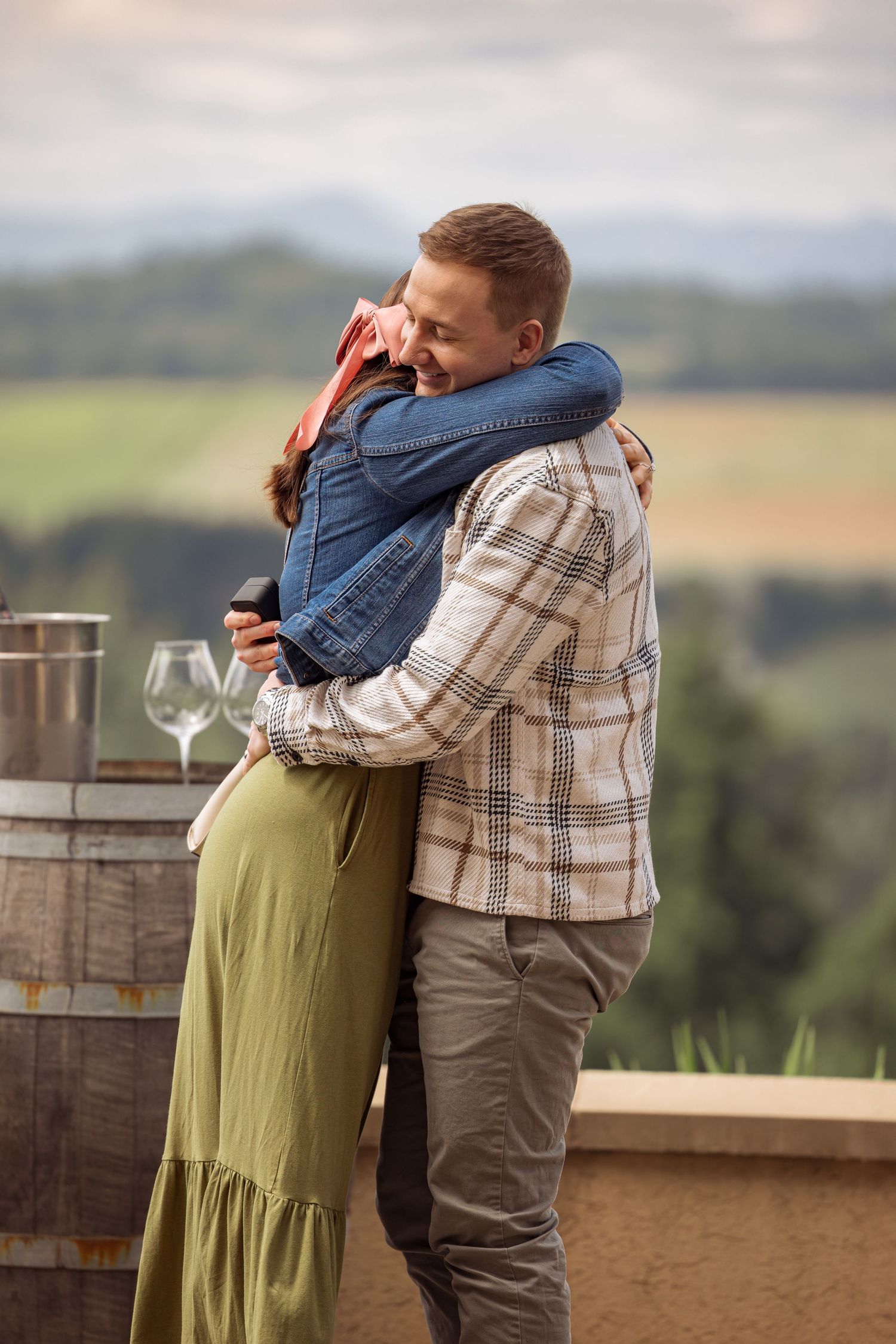 Couple embraces emotionally on a winery terrace overlooking scenic countryside with wine glasses nearby.