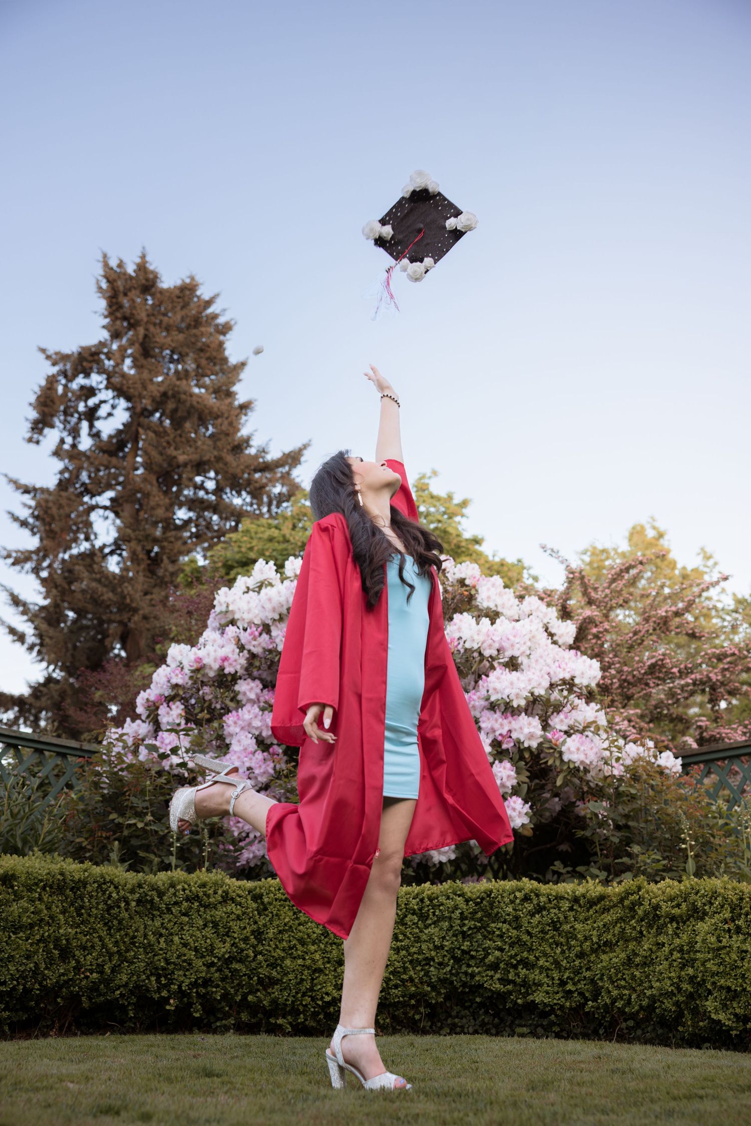 Young woman in red graduation gown tossing mortarboard cap in the air outdoors near blooming flowers.