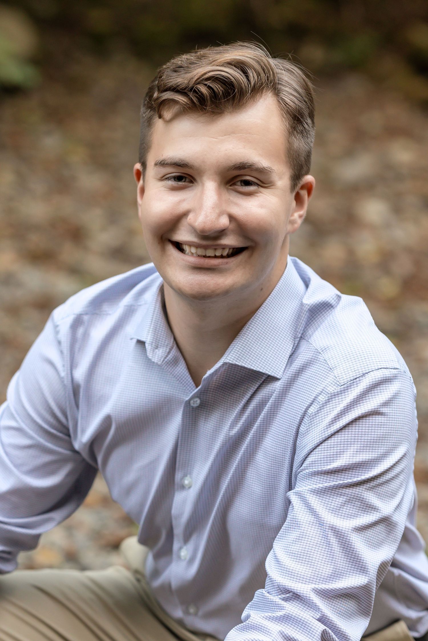 Professional headshot portrait of someone in a light blue button-down shirt smiling outdoors with blurred natural background.