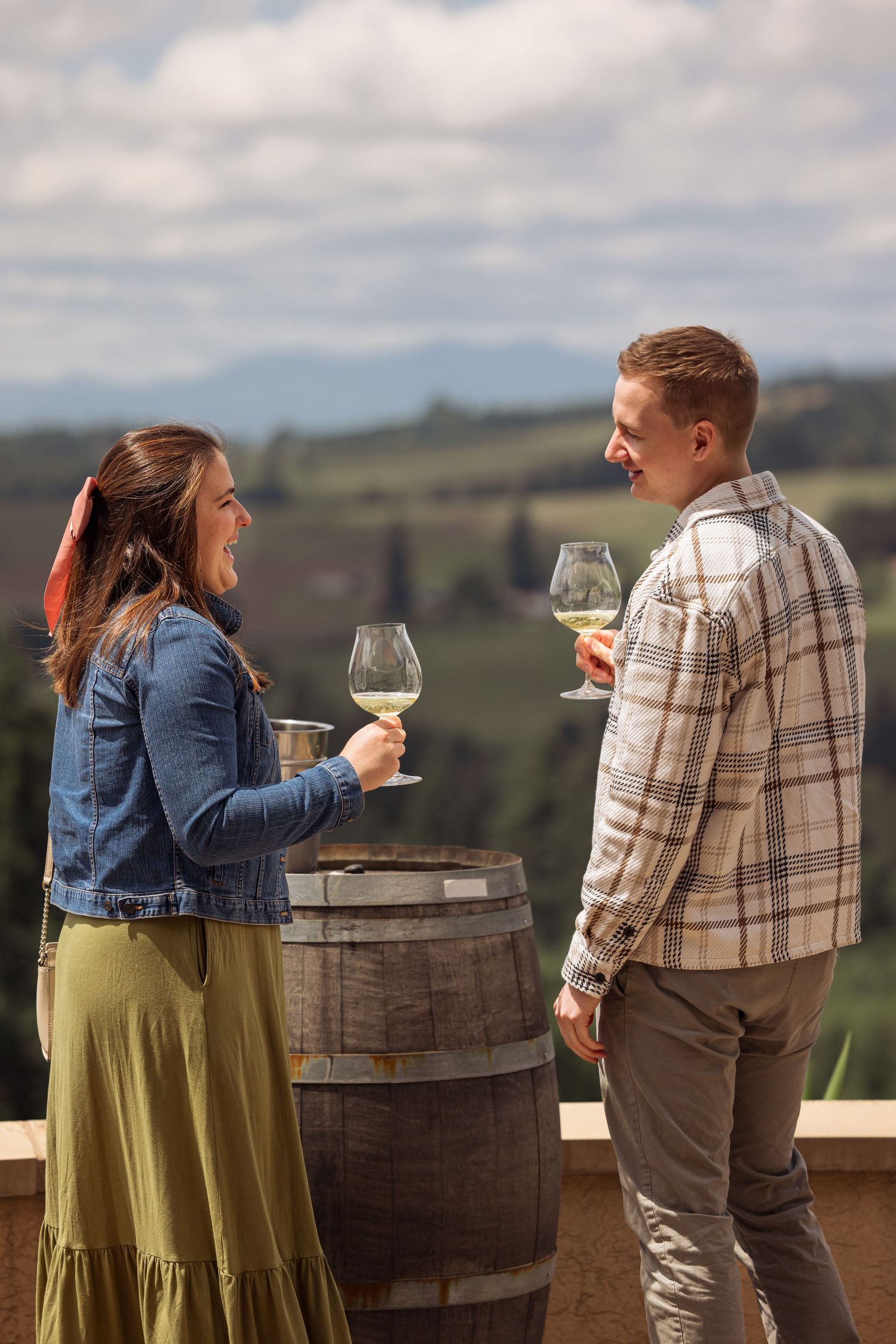 A couple standing by a wine barrel on a scenic hillside patio enjoying glasses of white wine.