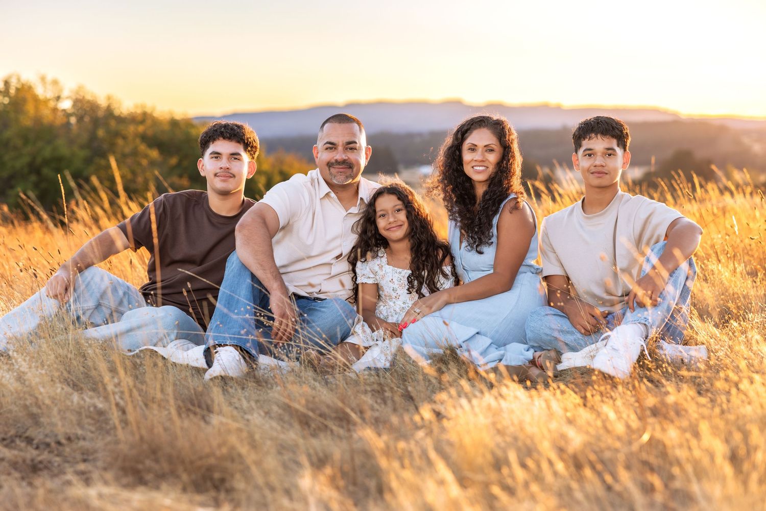 Family of five smiling together in golden field at sunset, sitting on blanket in warm evening light.