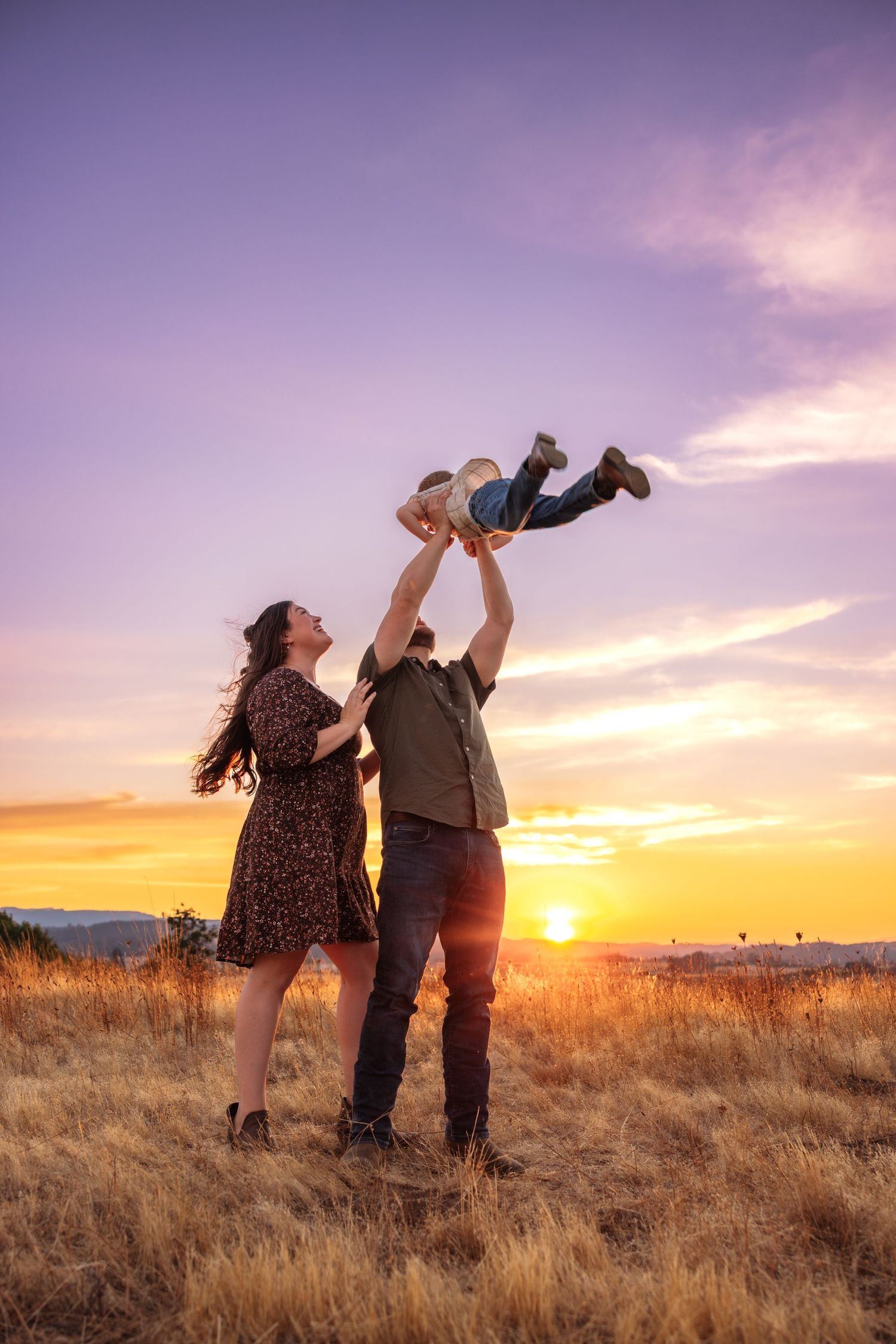 Family enjoying a golden sunset, father lifting child in the air while mother watches joyfully in a field.