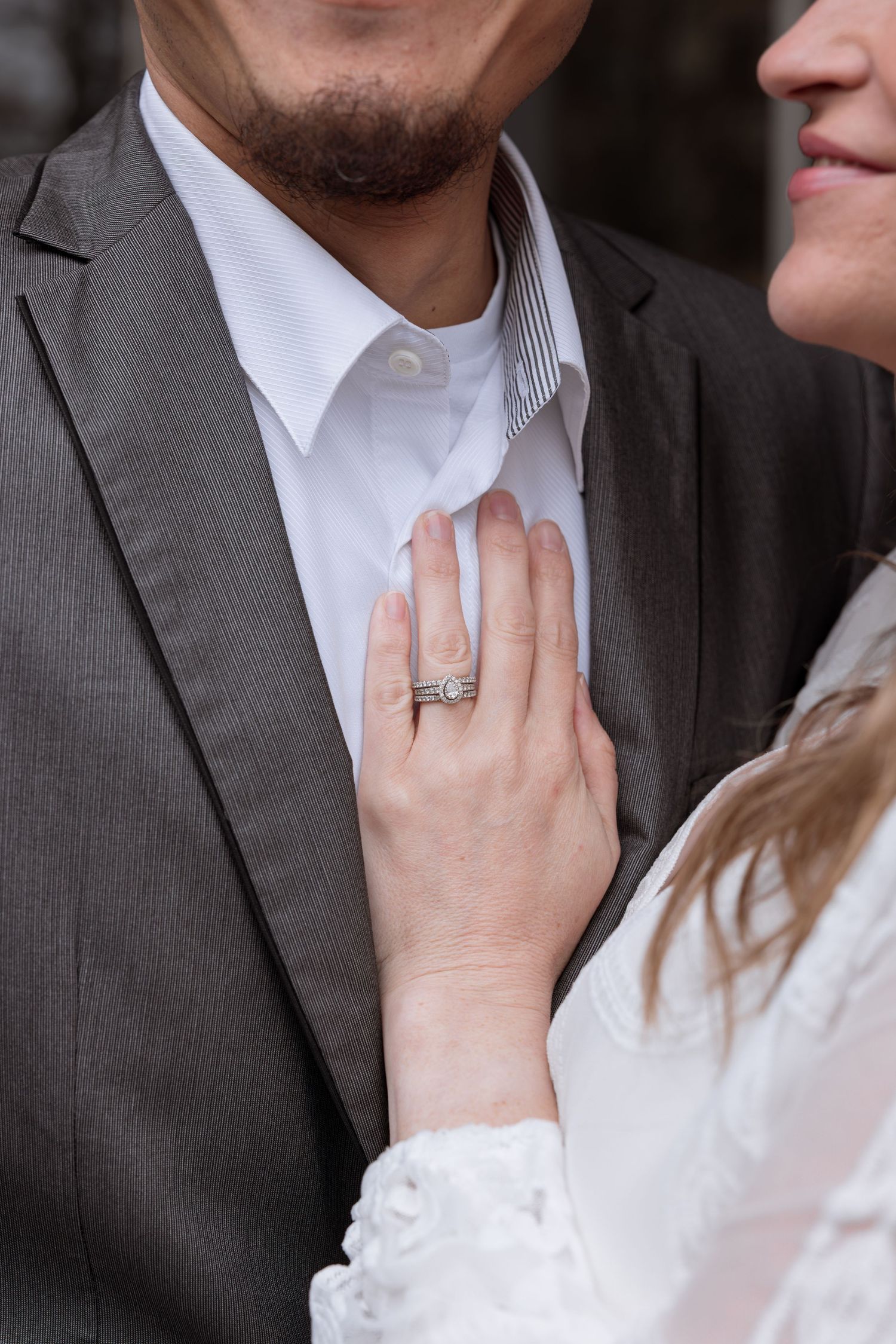 Close-up of a woman's hand with engagement ring resting on a man's chest in a gray suit.