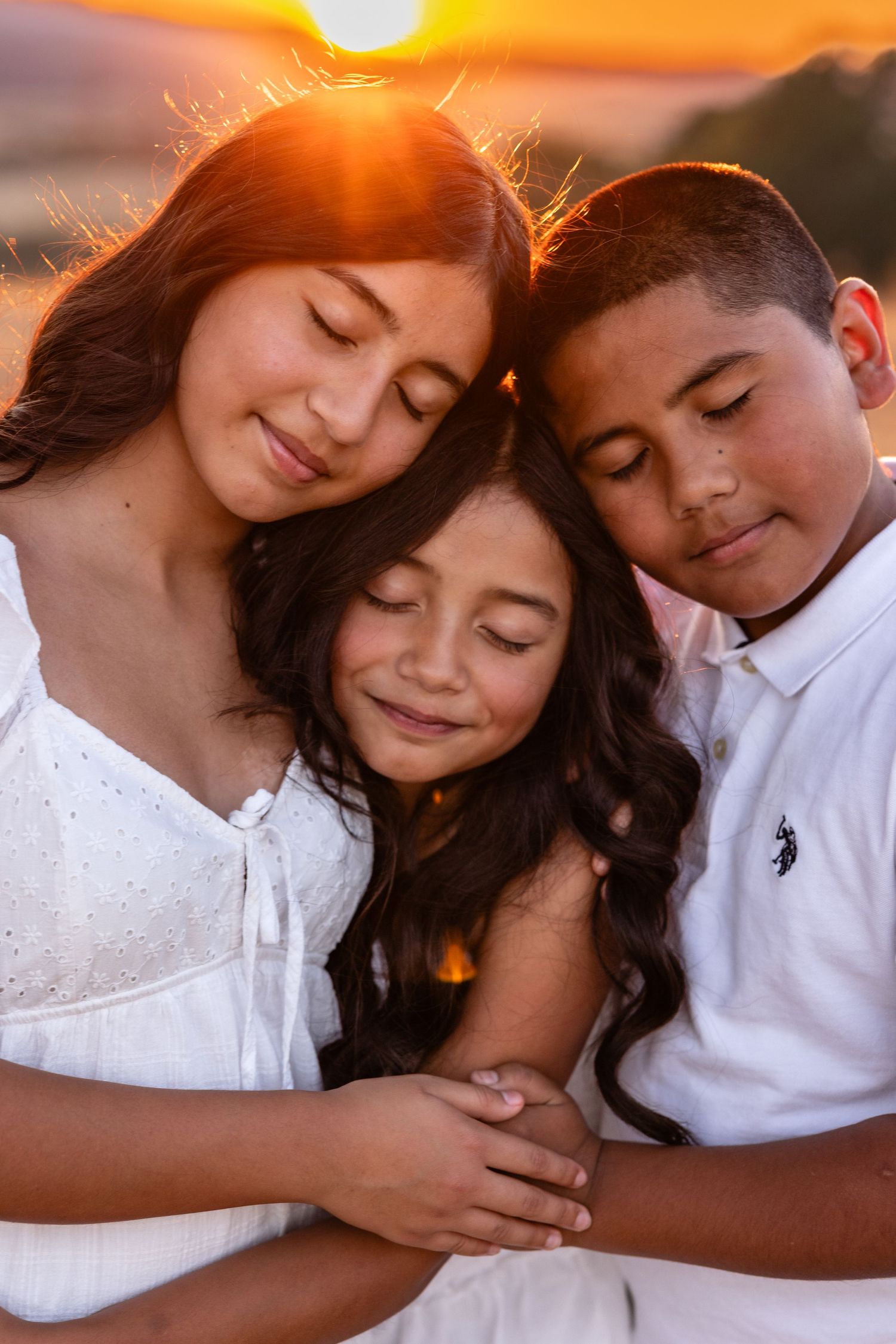 Three siblings share a tender embrace at golden sunset, eyes closed in peaceful moment, wearing white clothing outdoors.