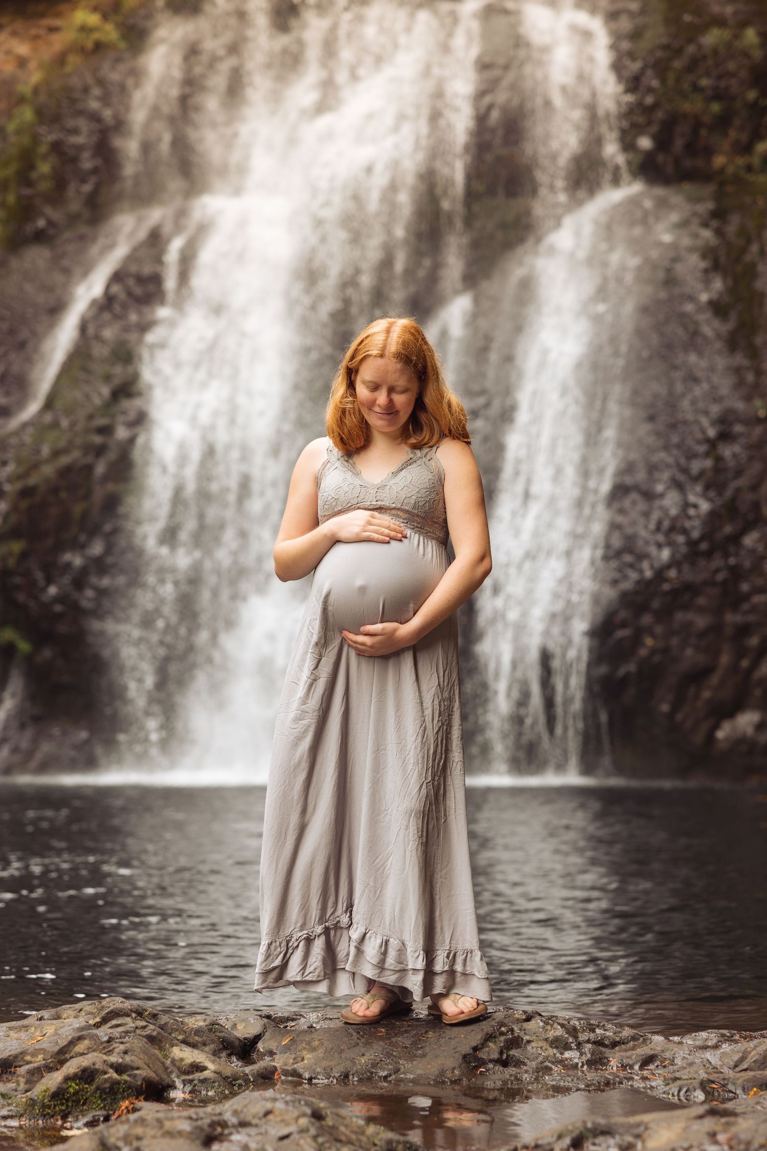 A pregnant woman in a flowy gray dress poses thoughtfully by a cascading waterfall in a serene natural setting.