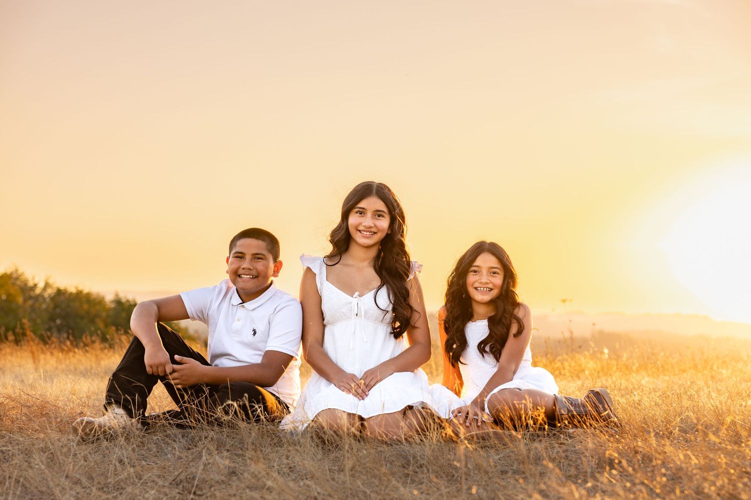 Three children in white outfits sitting in golden field at sunset, smiling warmly in a beautiful outdoor portrait.