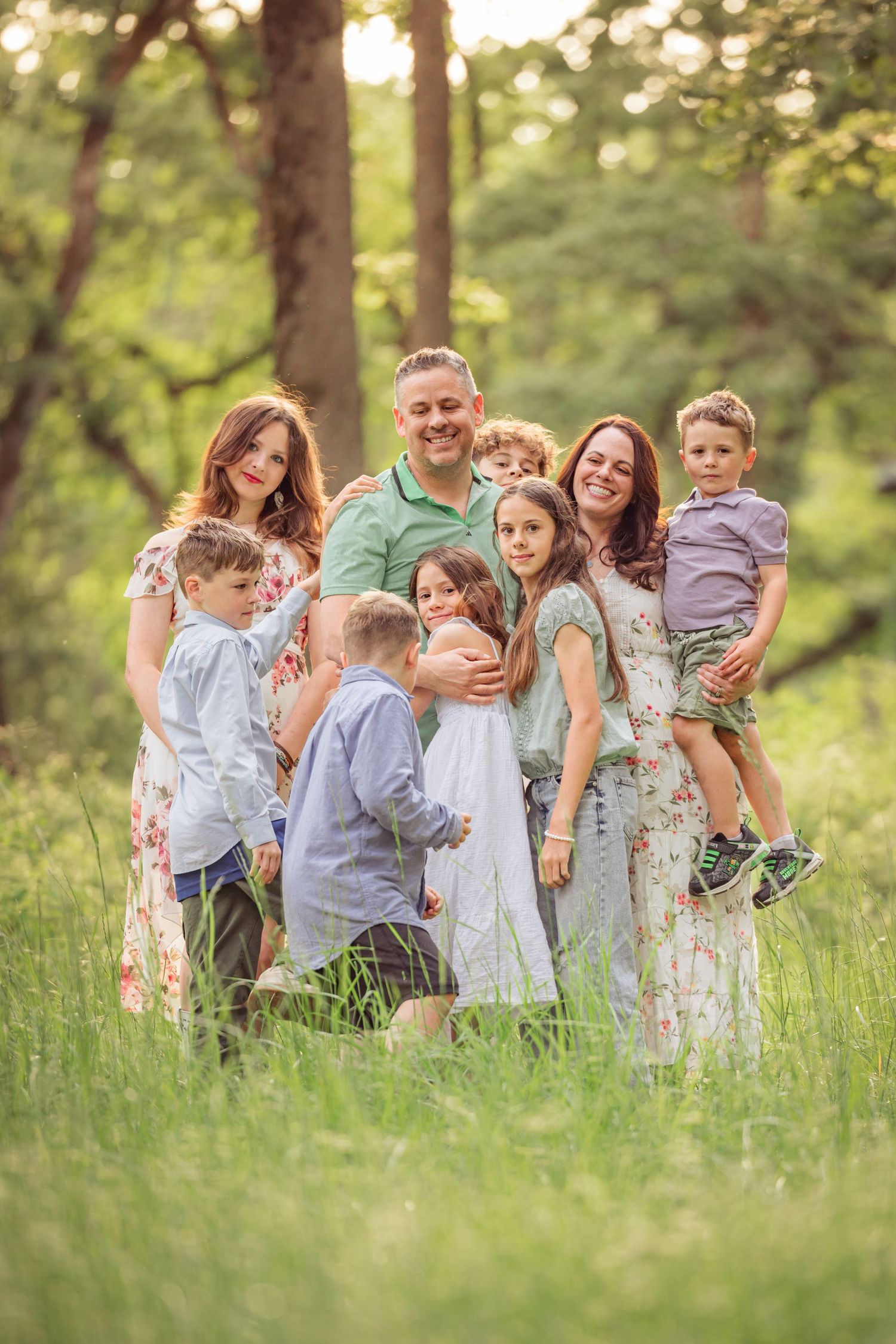 A family poses together outdoors in tall grass with trees in the background during golden hour lighting.
