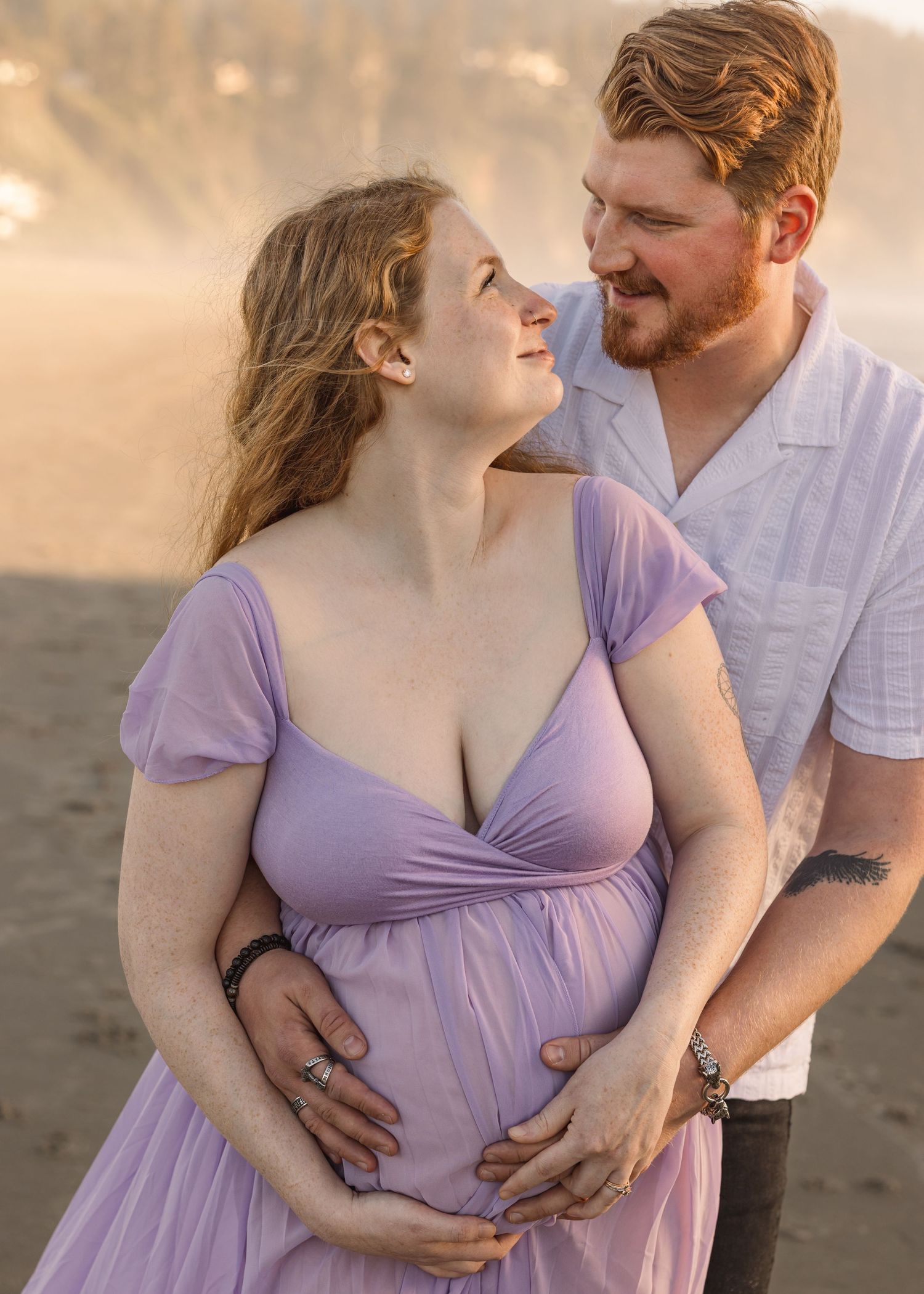 A couple shares an intimate moment on the beach at sunset, with the person in purple wearing a flowing dress.