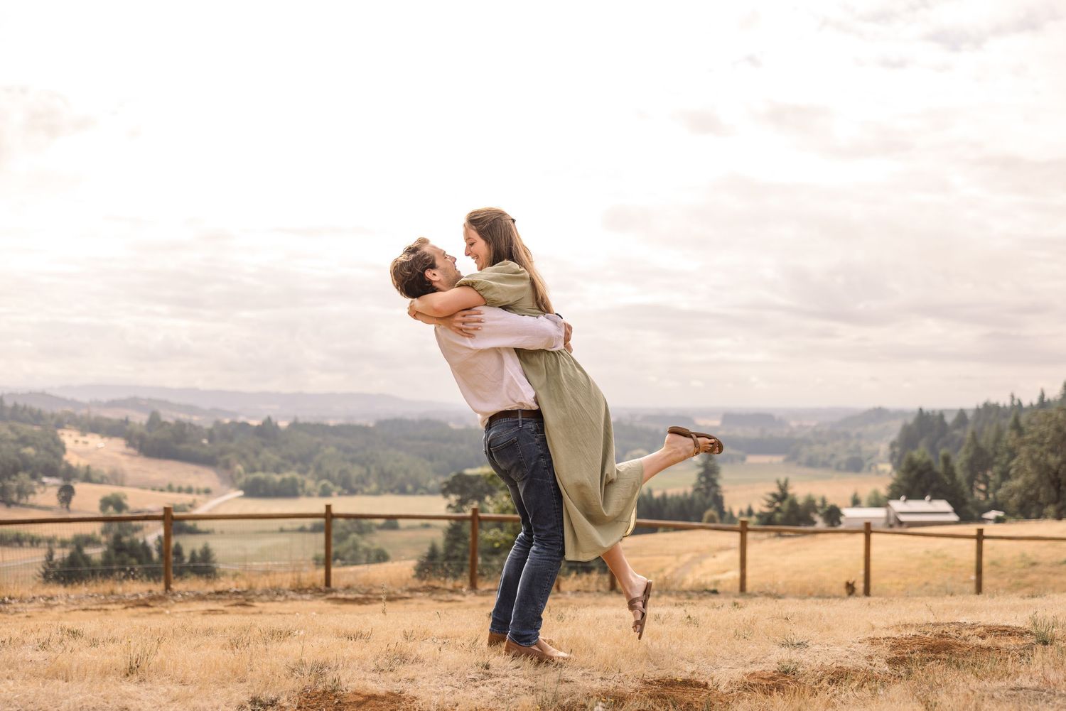 A couple embraces while standing on a wooden fence overlooking rolling hills and countryside at sunset.
