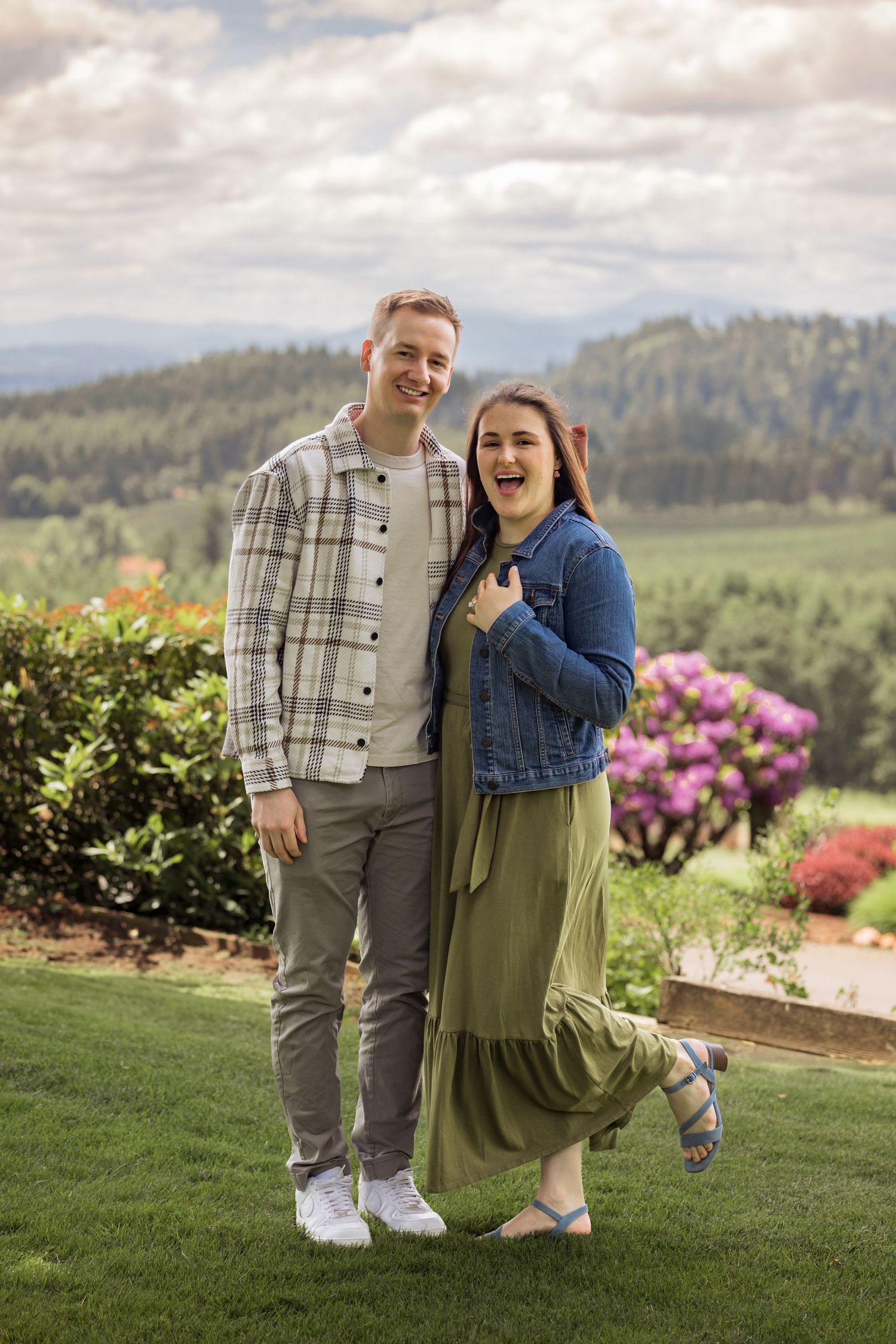 A couple shares a joyful moment on a grassy hilltop with blooming flowers and mountain views in the background.