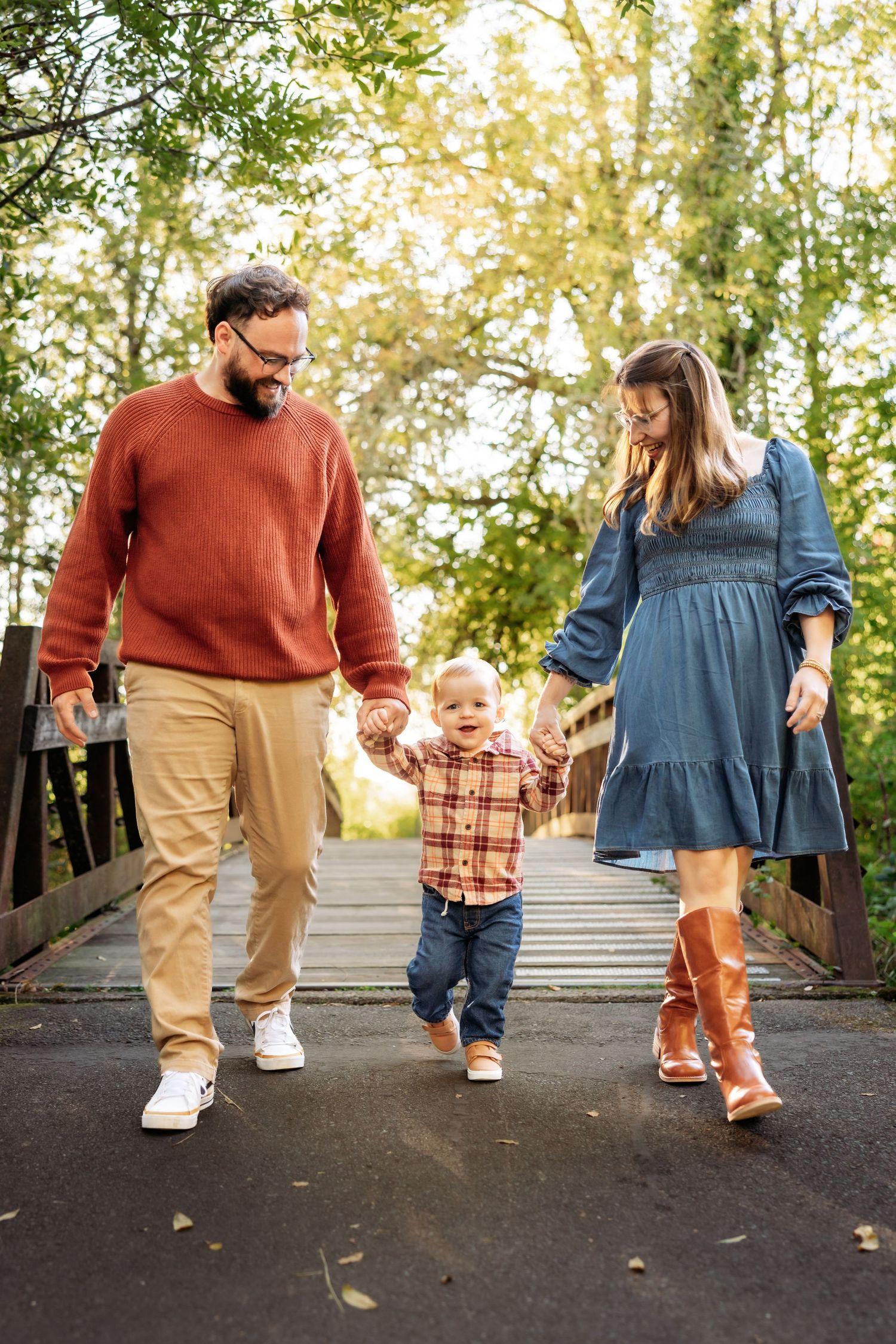 A happy family with a toddler walks hand-in-hand on a bridge surrounded by autumn trees.