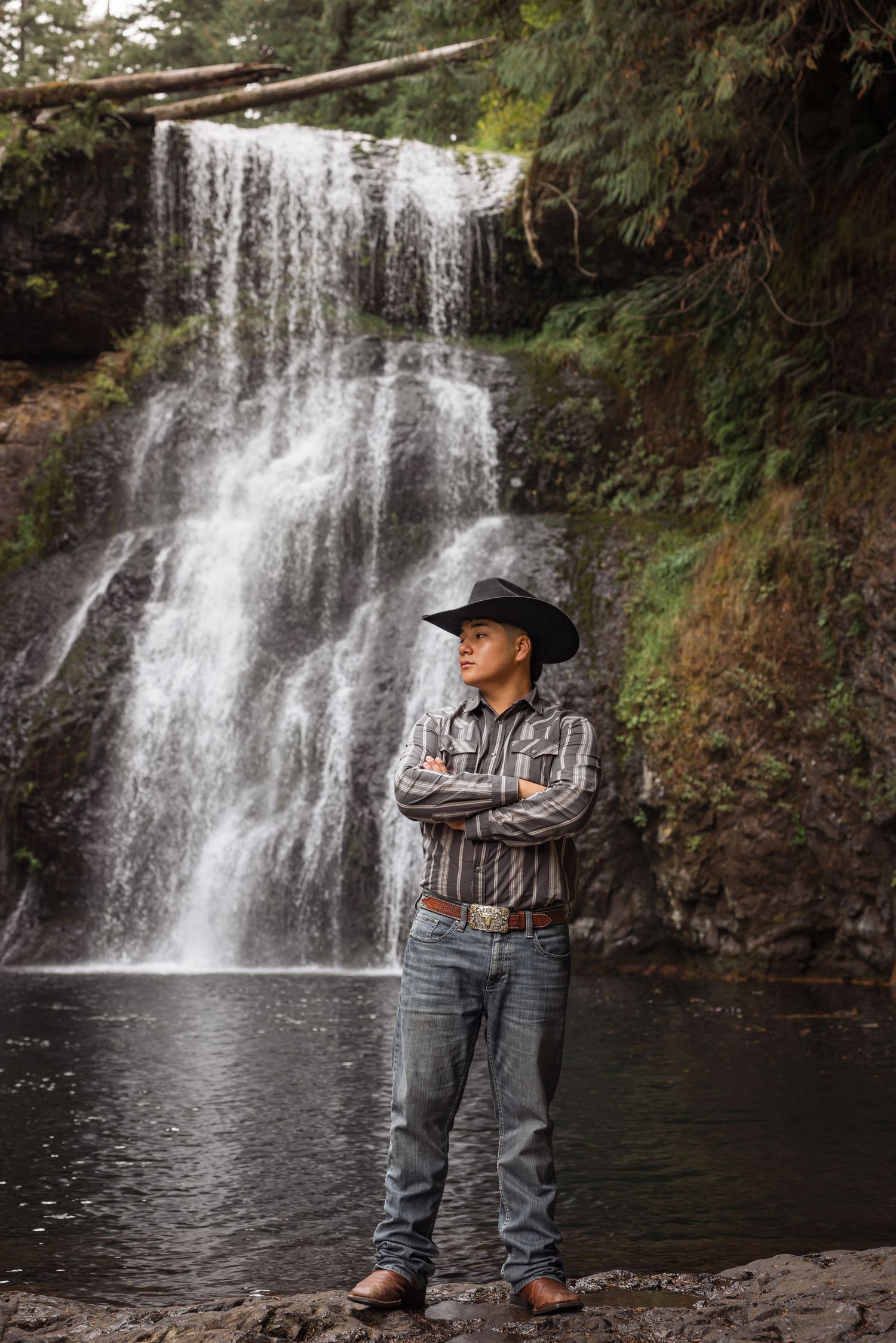 Man in cowboy hat and plaid shirt stands arms crossed before a lush forest waterfall.