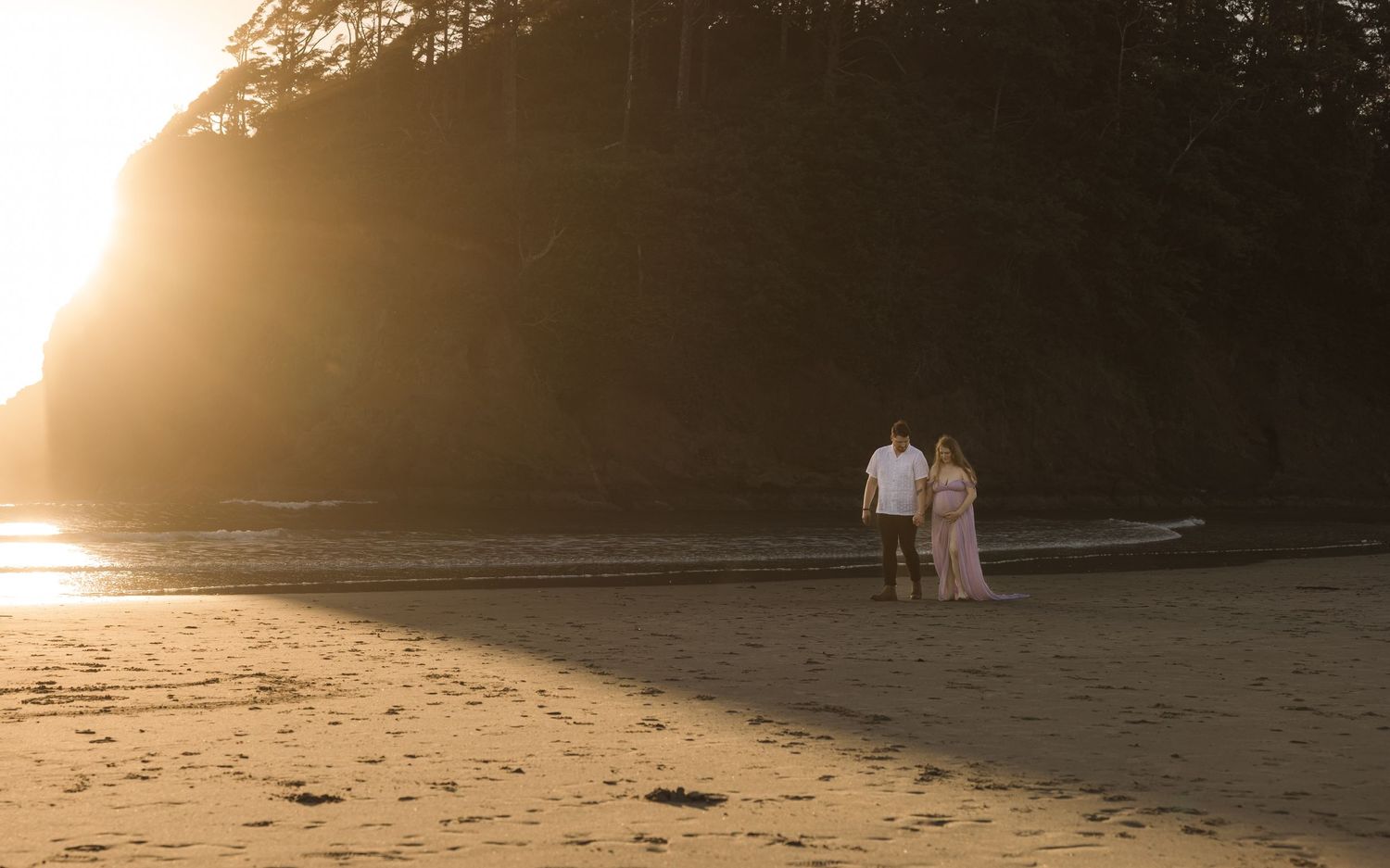 Two figures walk along a serene beach at sunset with golden light streaming through trees in the background.