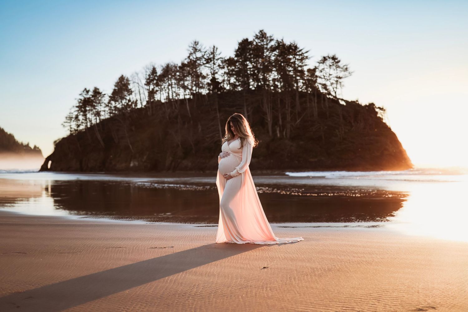 Scenic maternity photo on beach at sunset with dramatic rock formation in background.