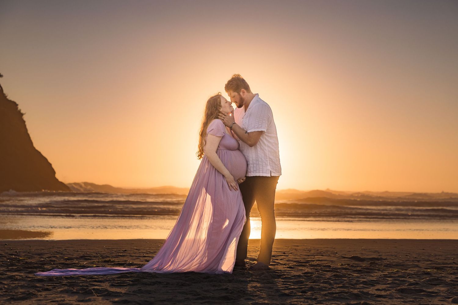 A couple embraces on a beach at sunset with golden light and crashing waves in the background.