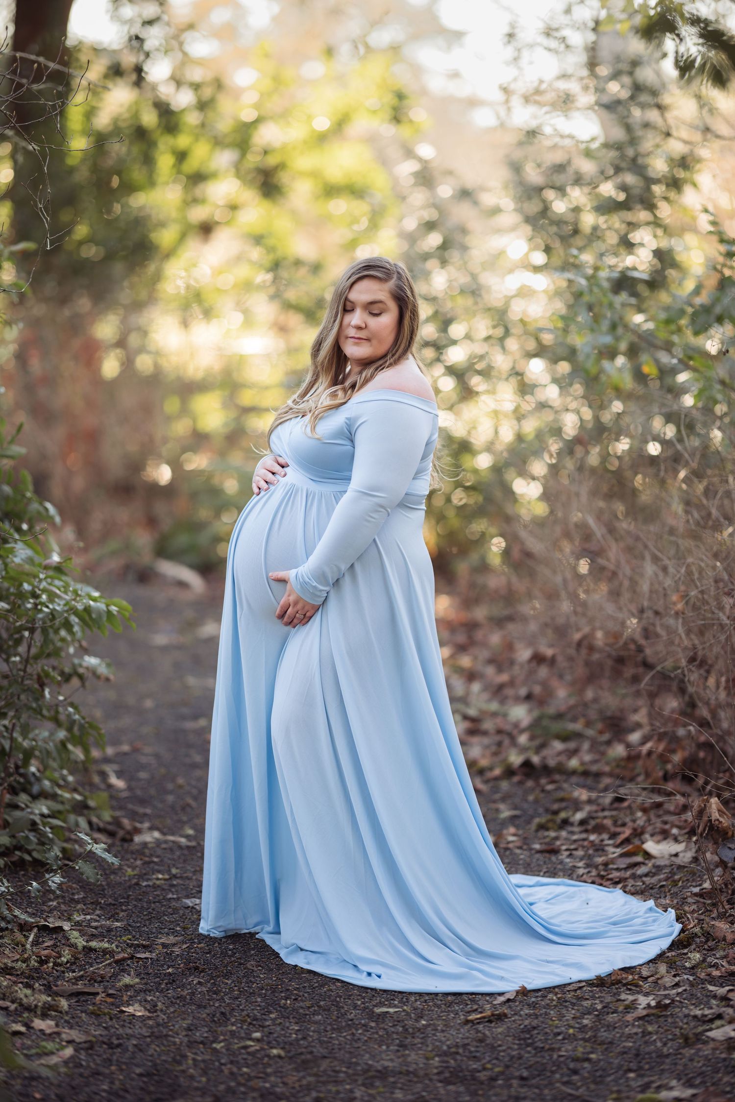 A maternity photoshoot in a forest setting features a flowing light blue gown with a long train against natural foliage.