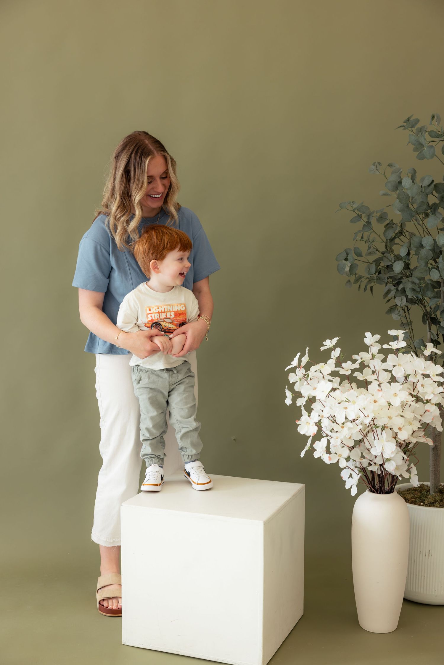A series of studio photos showing a child in light blue denim and white sneakers posing on a white box near floral decor.