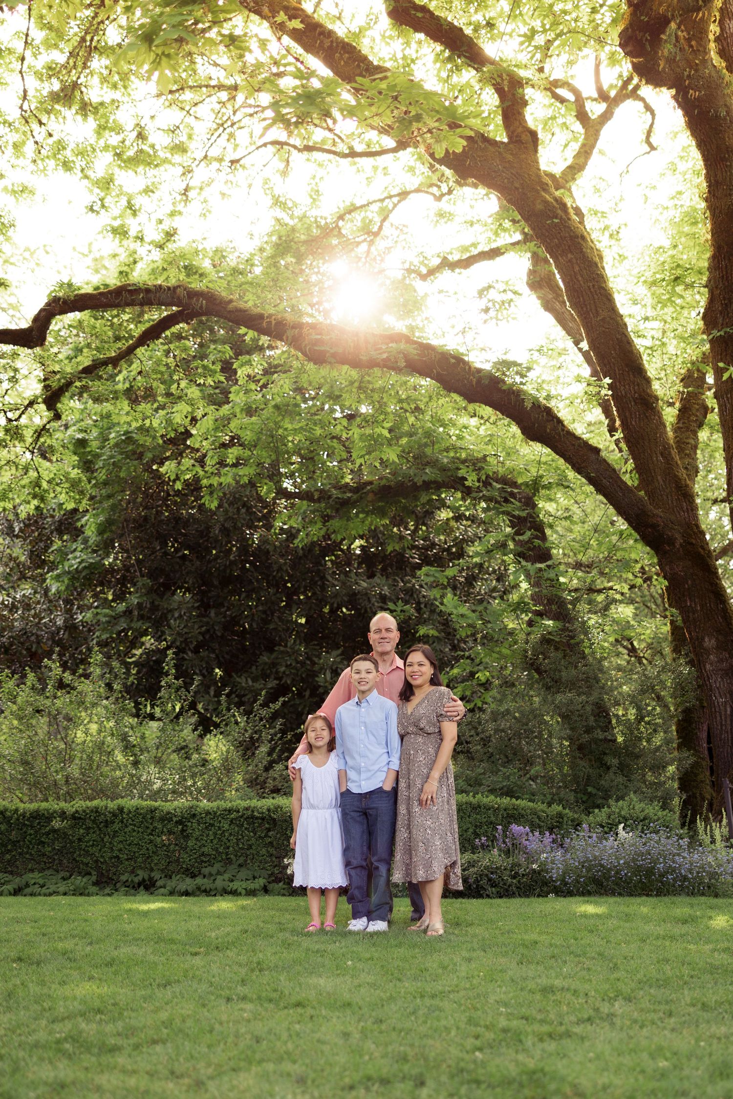 Family portrait under a majestic tree with sunlight streaming through branches in garden setting.
