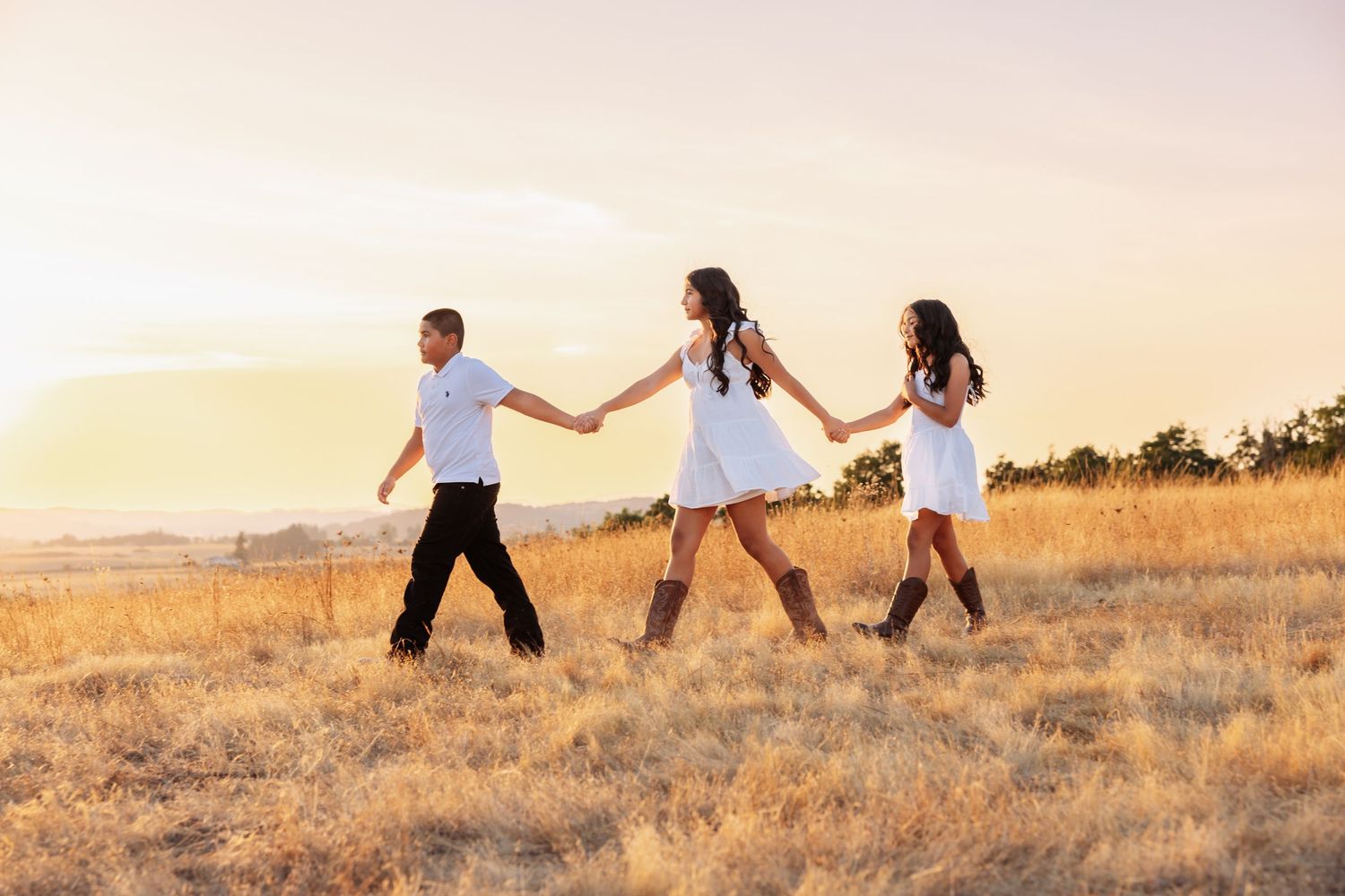 A man and two women in white dresses and cowboy boots hold hands walking through a golden field at sunset.